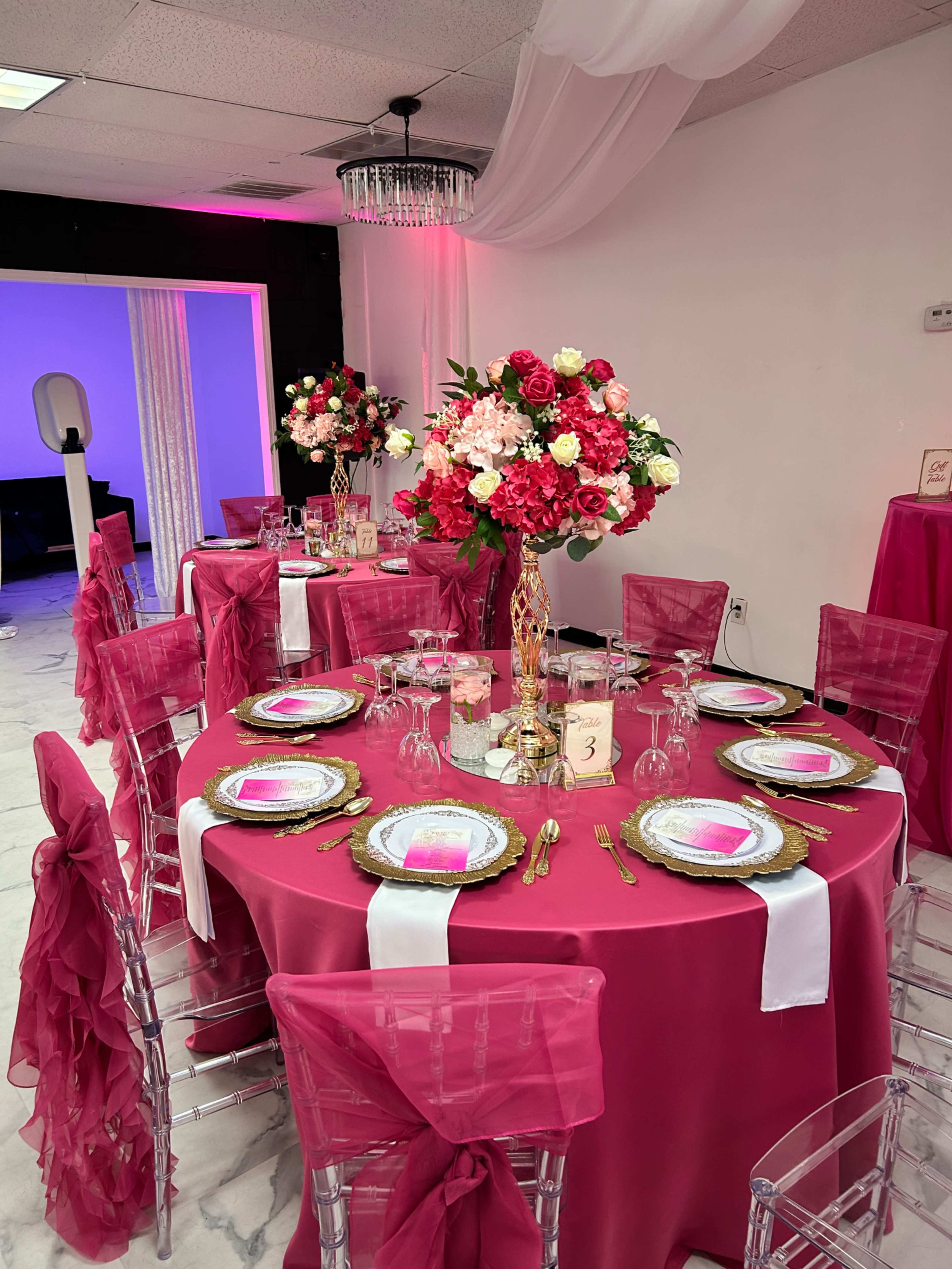 The image shows a banquet setup with round tables dressed in pink tablecloths, adorned with floral centerpieces, elegant tableware, and decorative seating arrangements.