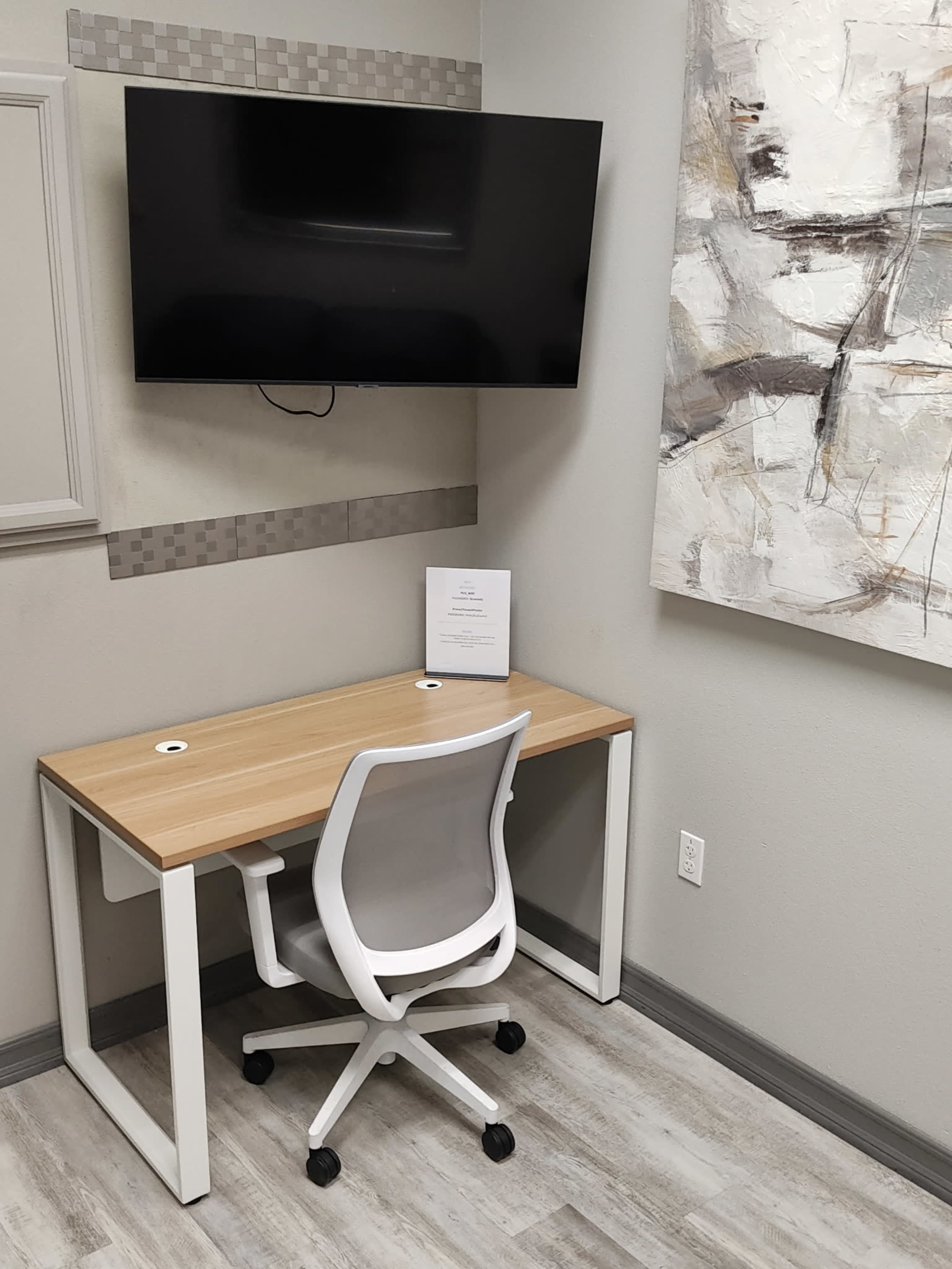 The image shows a minimalist workspace featuring a wooden desk with a white chair, a mounted television on the wall, and an abstract painting nearby.