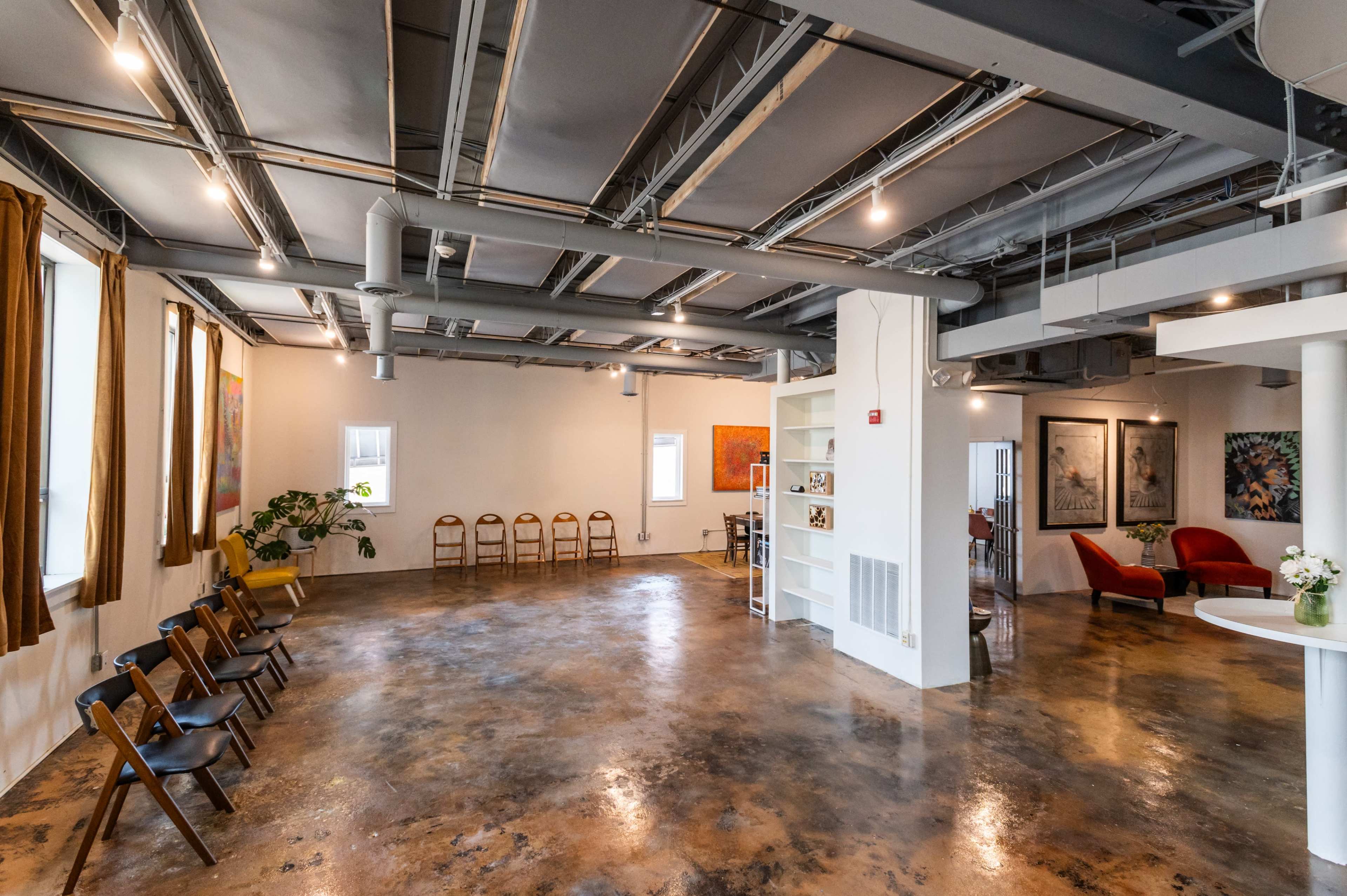 A spacious indoor area with a polished concrete floor, featuring several chairs arranged along the walls and artwork hanging on the white walls.
