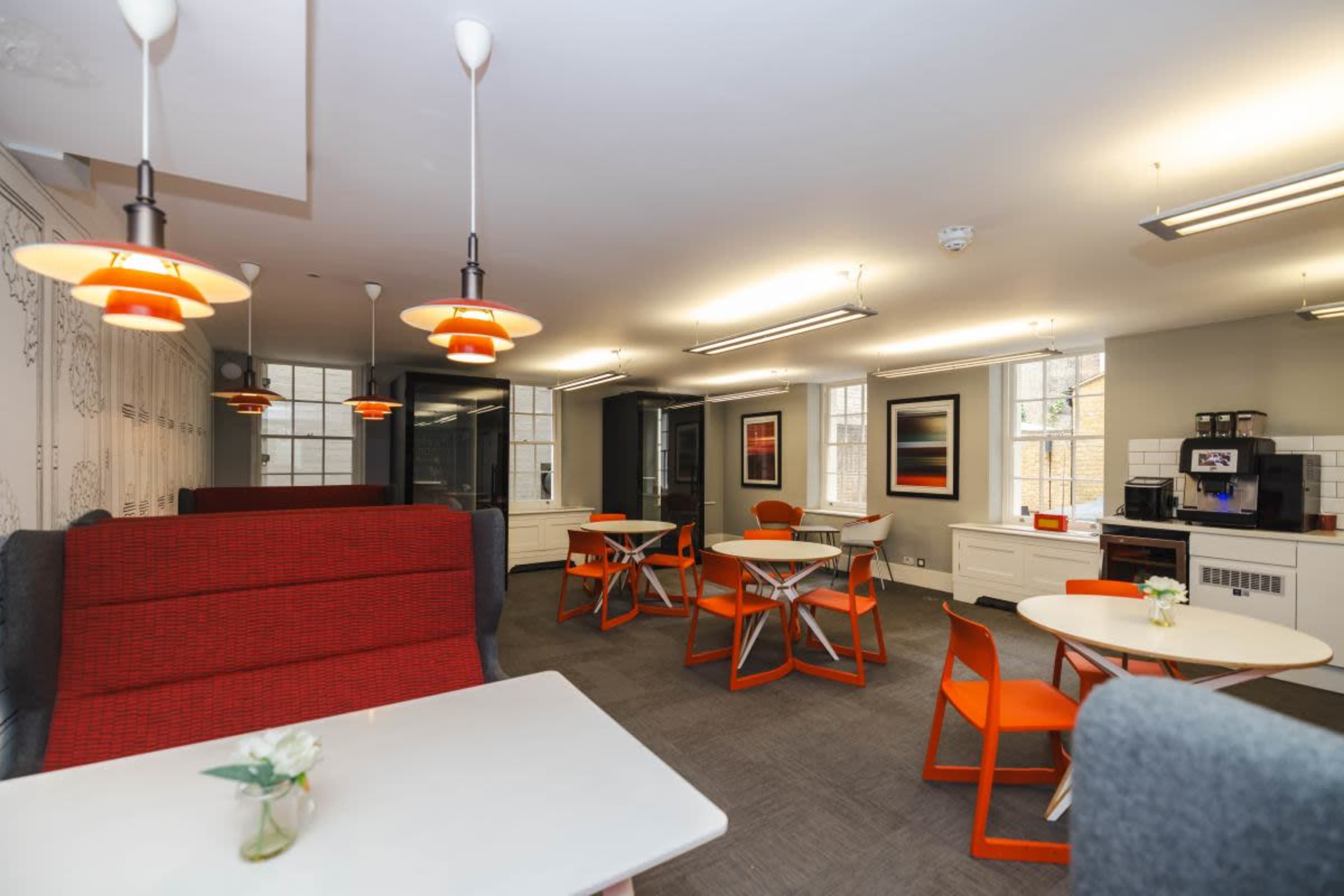 The image shows a modern lounge area with orange and gray furniture, featuring tables and chairs arranged for dining or gathering, and bright lighting.