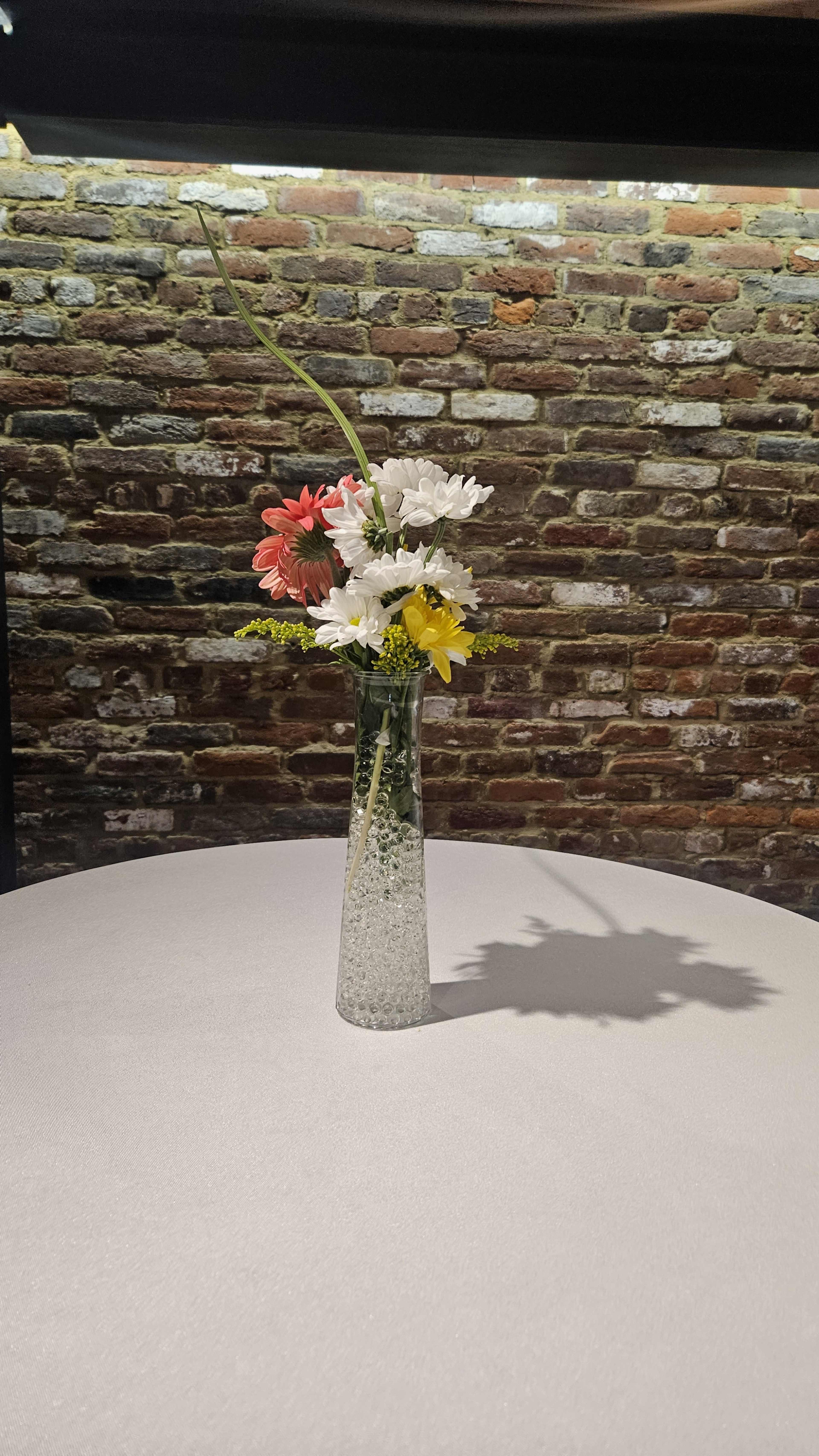 A glass vase filled with flowers stands on a round, white table in front of a brick wall.