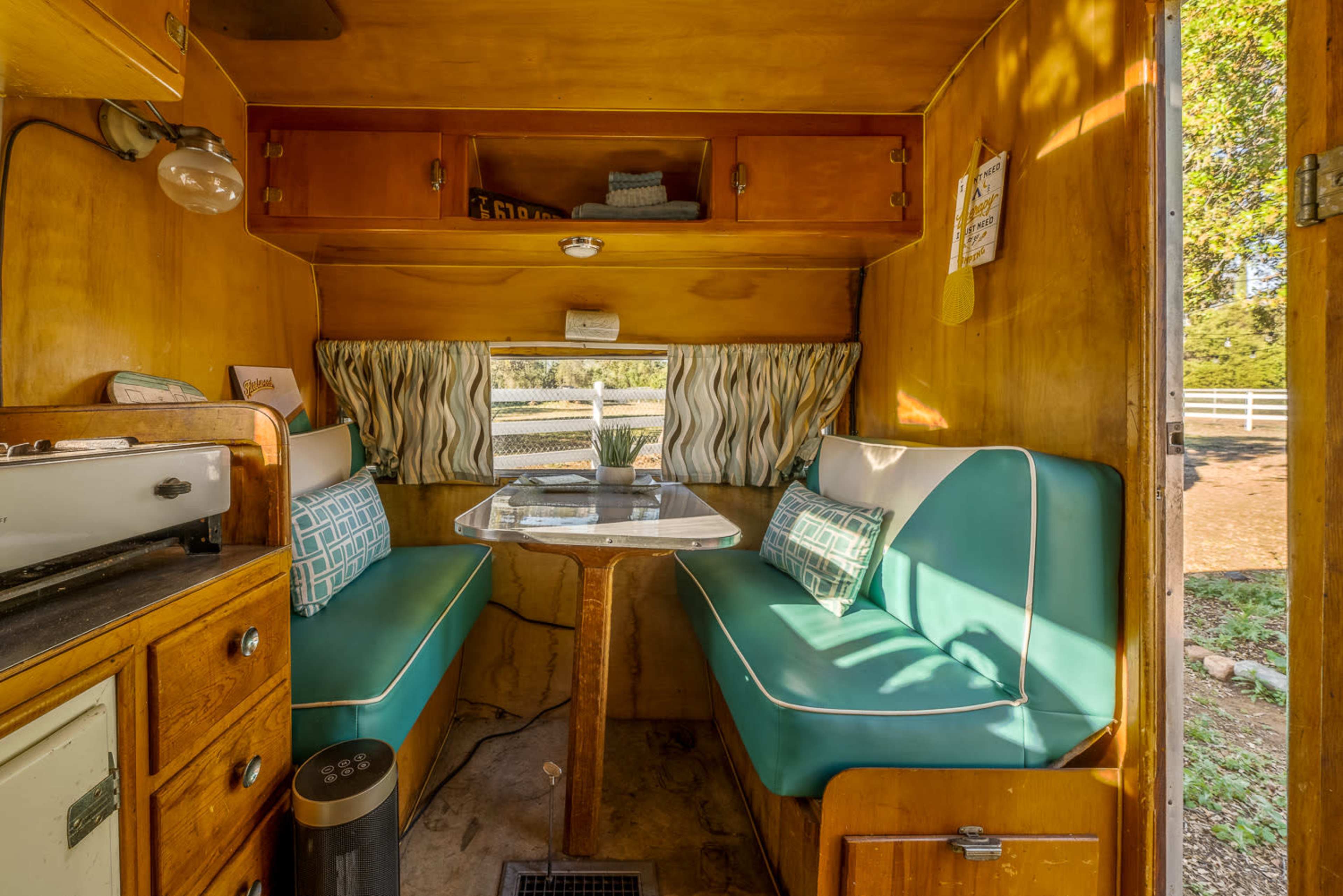 The interior of a vintage camper features a small dining table surrounded by teal and cream cushioned benches with wooden cabinetry and windows letting in natural light.