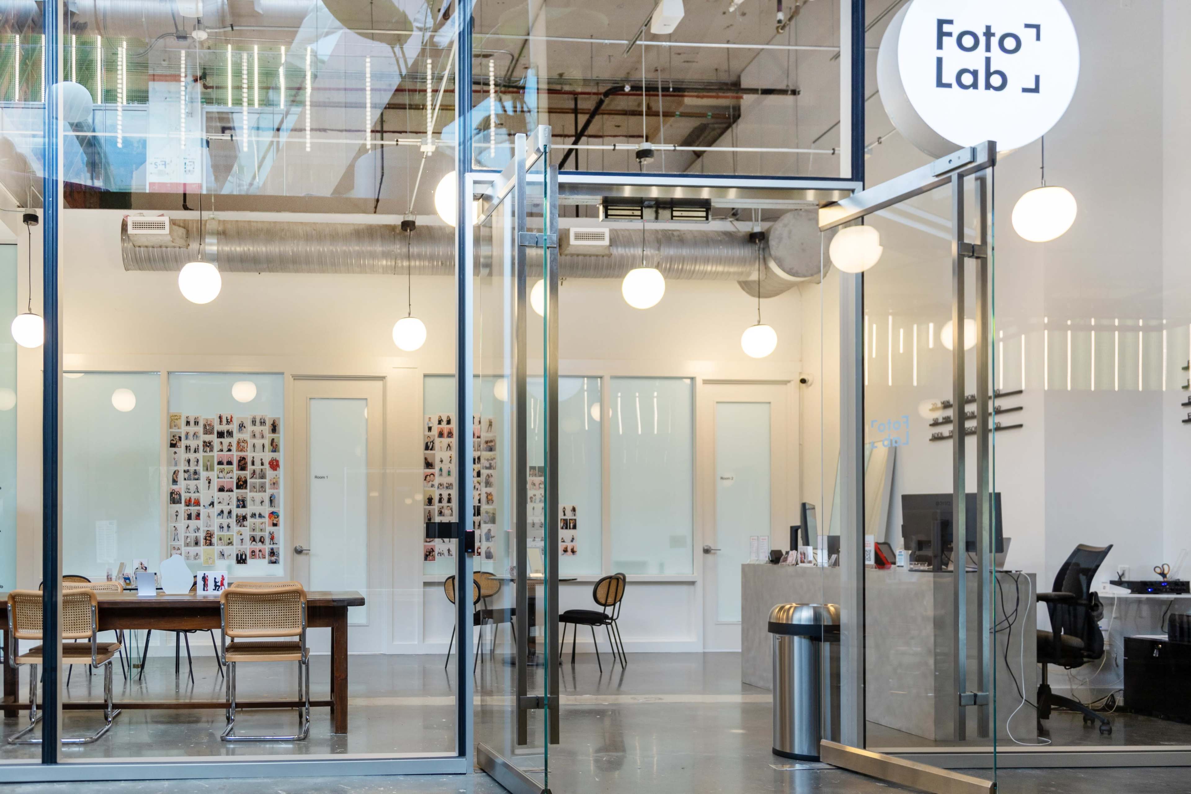 The entrance of a modern photo lab with large glass doors, a long table, and a wall adorned with photo prints.