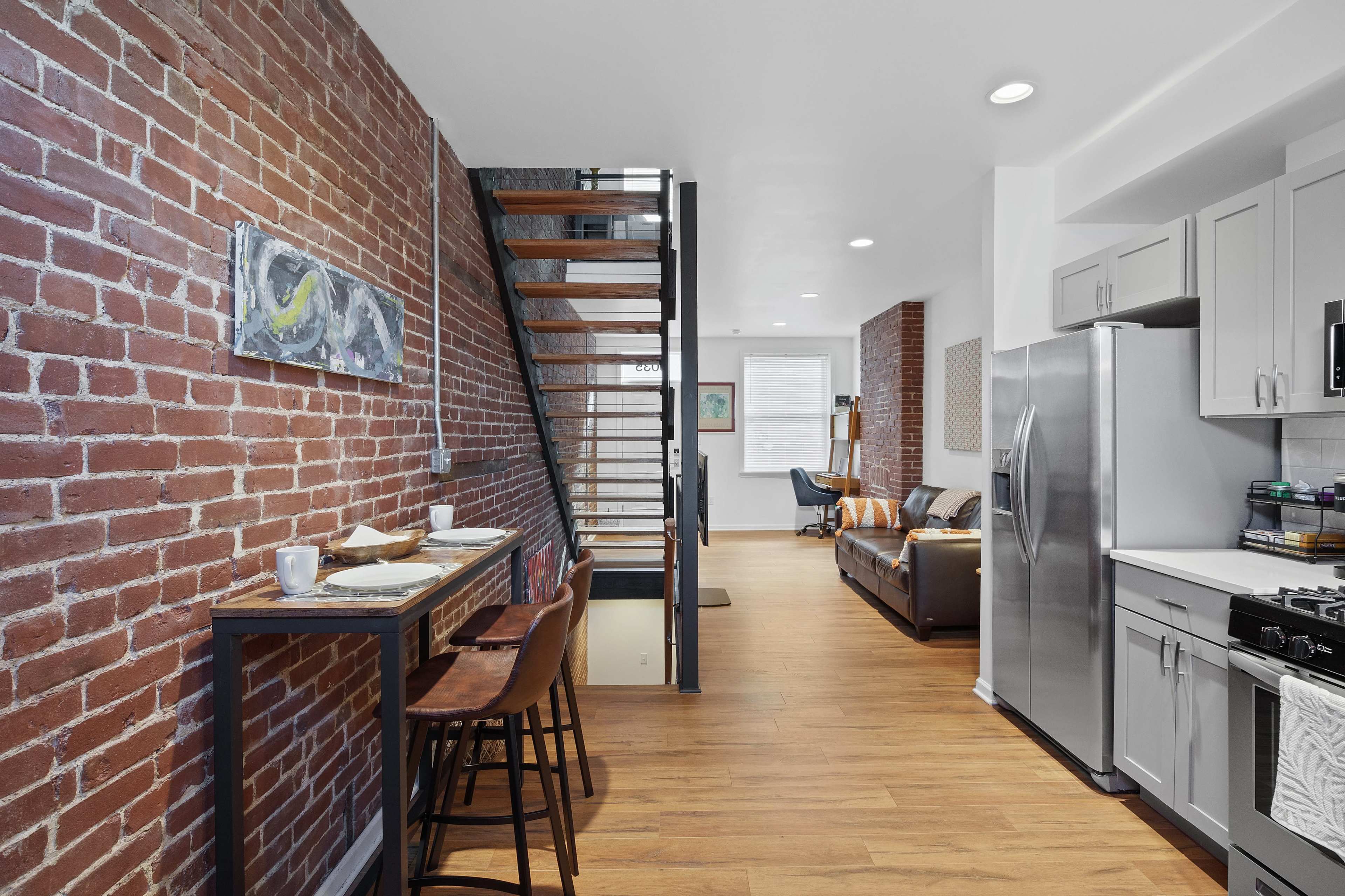 The image shows a modern kitchen and living area featuring exposed brick walls, a staircase, and contemporary furniture.
