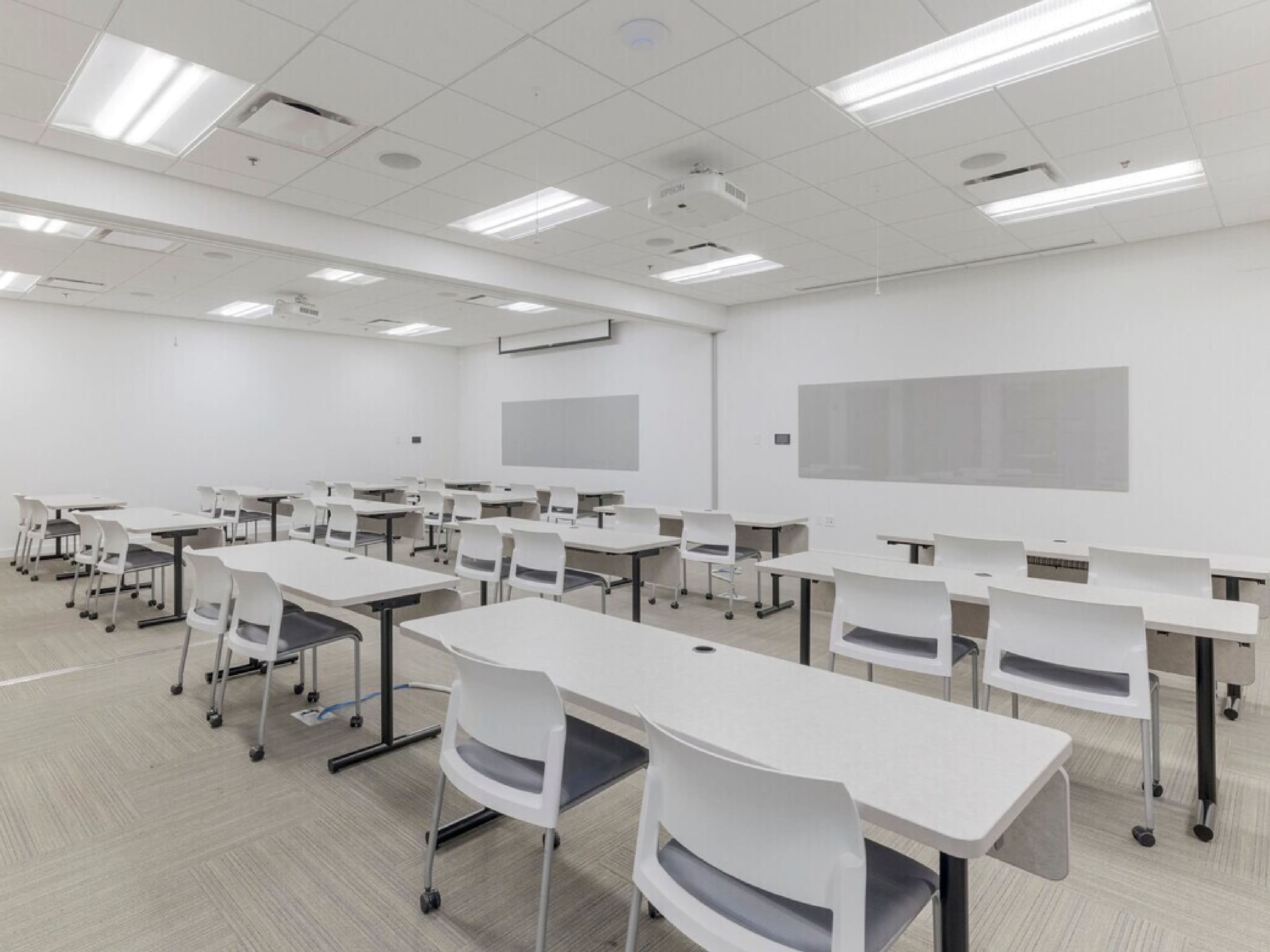 The image shows a sparsely furnished classroom with multiple tables and chairs arranged in rows, and blank walls with a projector mounted on the ceiling.
