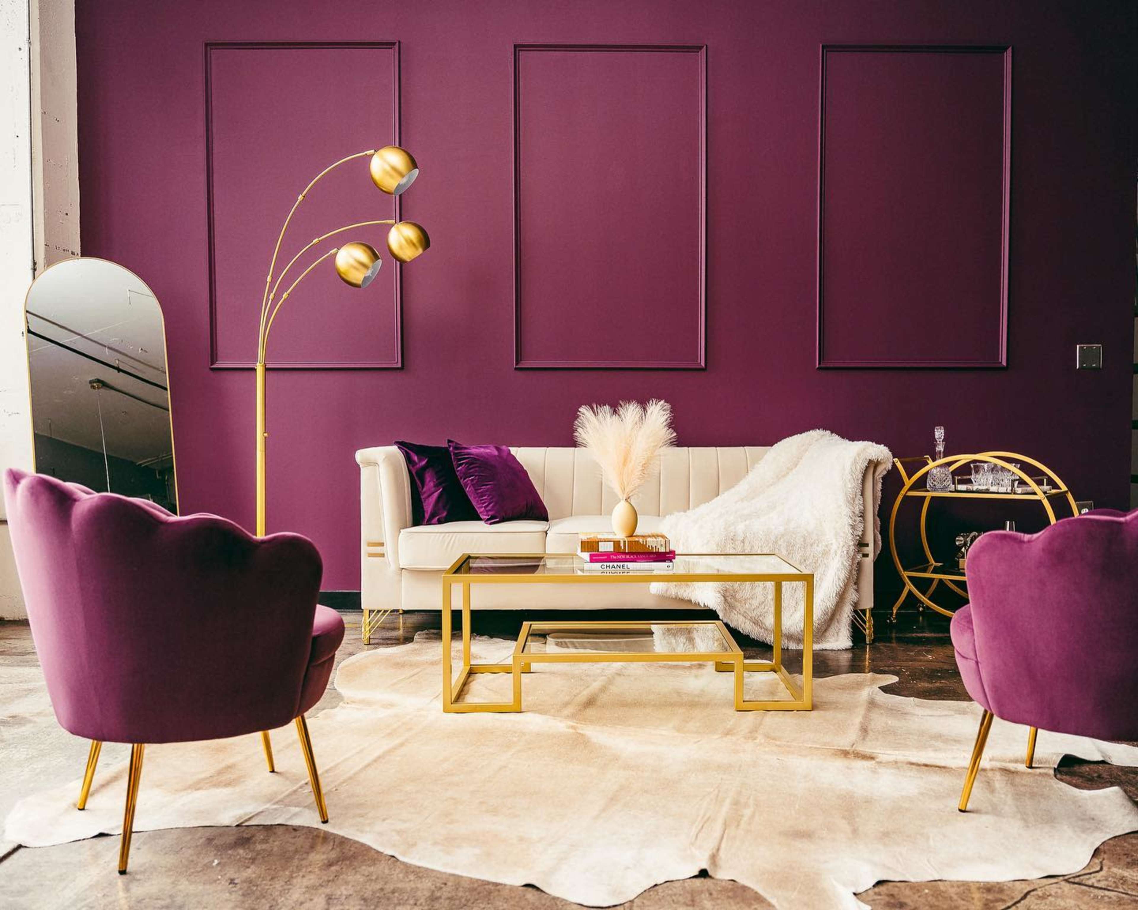 A modern living room features a white sofa, two purple armchairs, a gold coffee table, and a floor lamp against a dark purple wall.