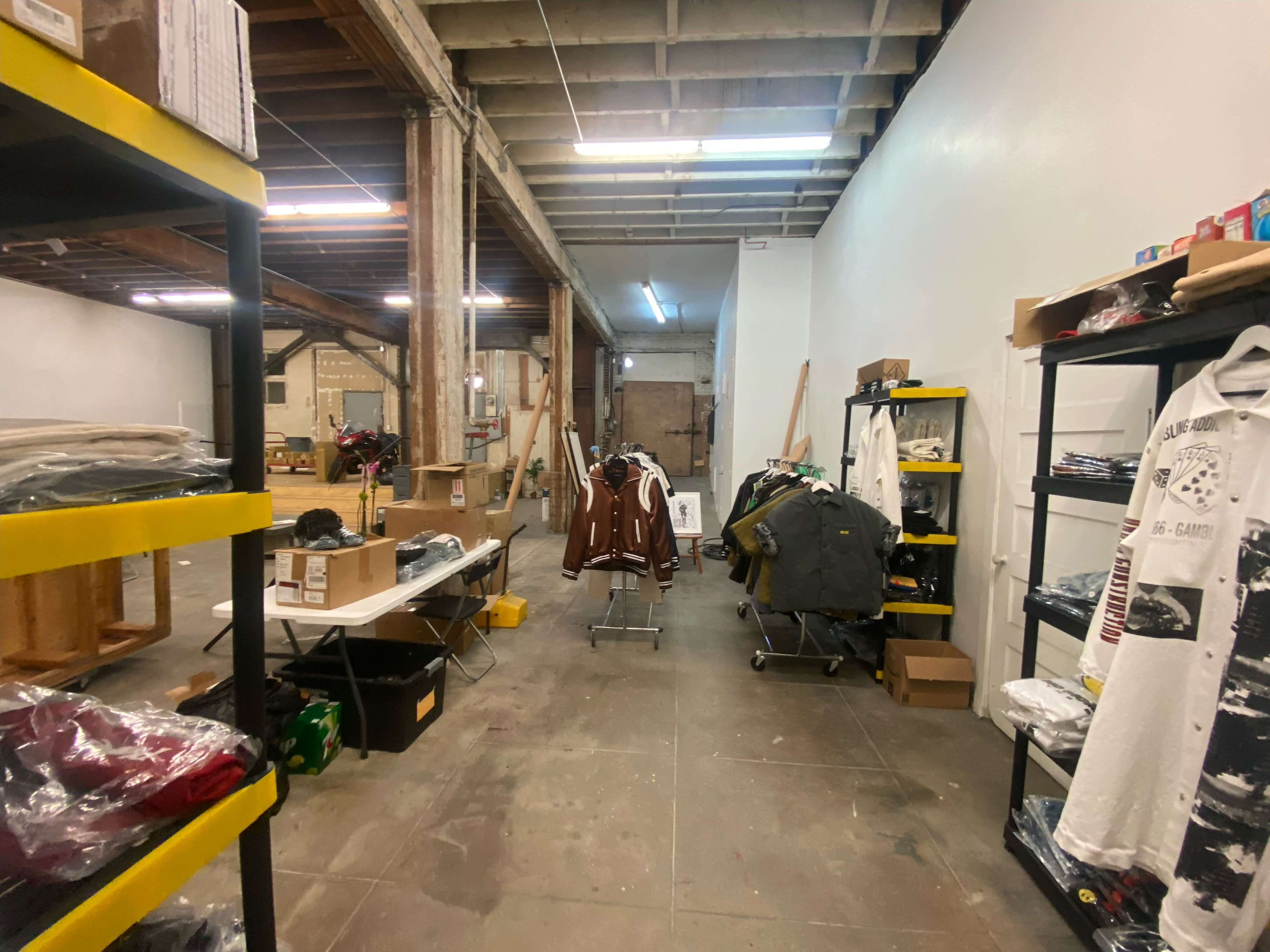 A large, empty workspace featuring shelves filled with clothing, tables with cardboard boxes, and a clothing rack in a well-lit environment.