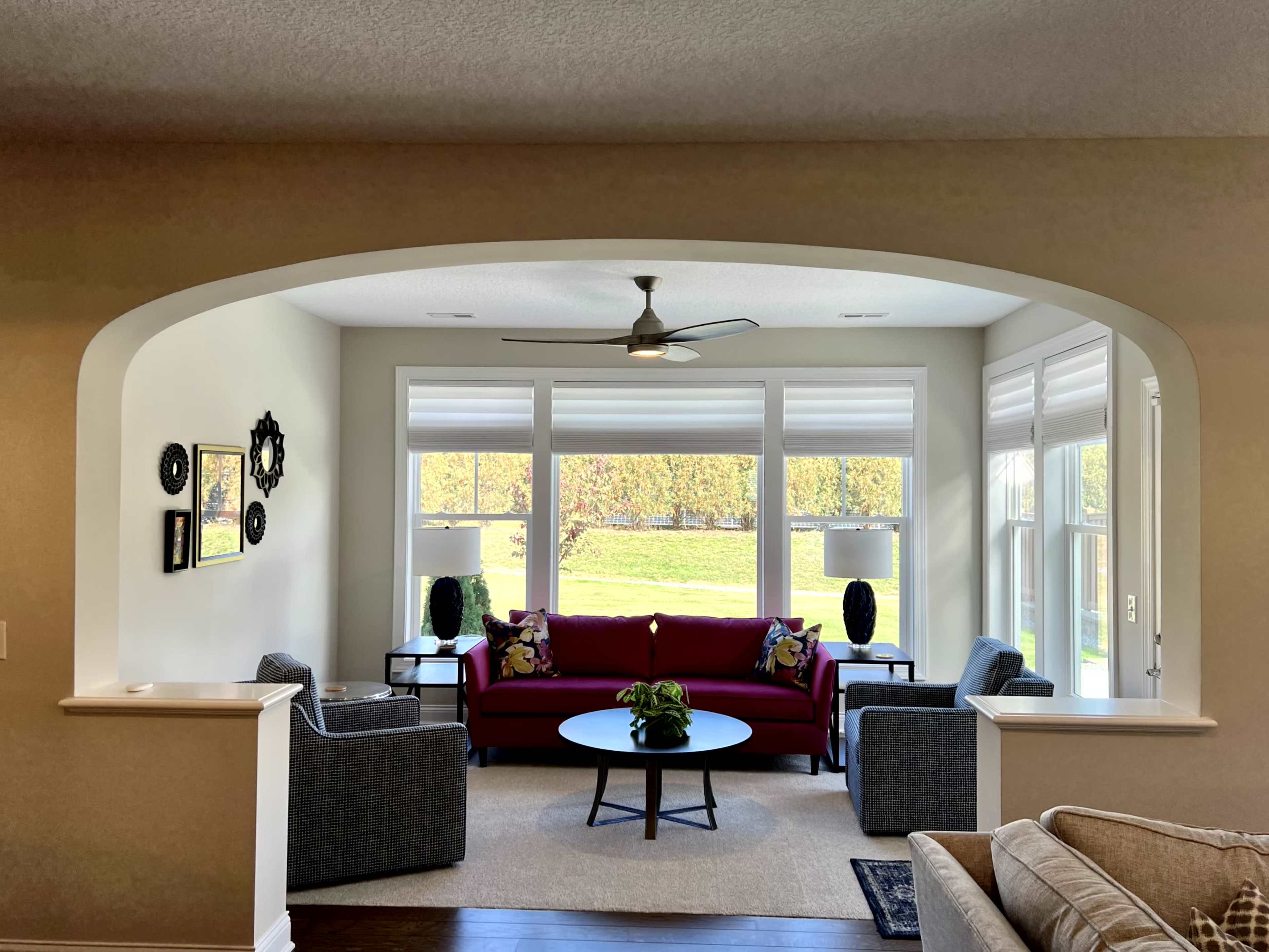 The image shows a living room with a red sofa, gray armchairs, and large windows overlooking a green landscape.