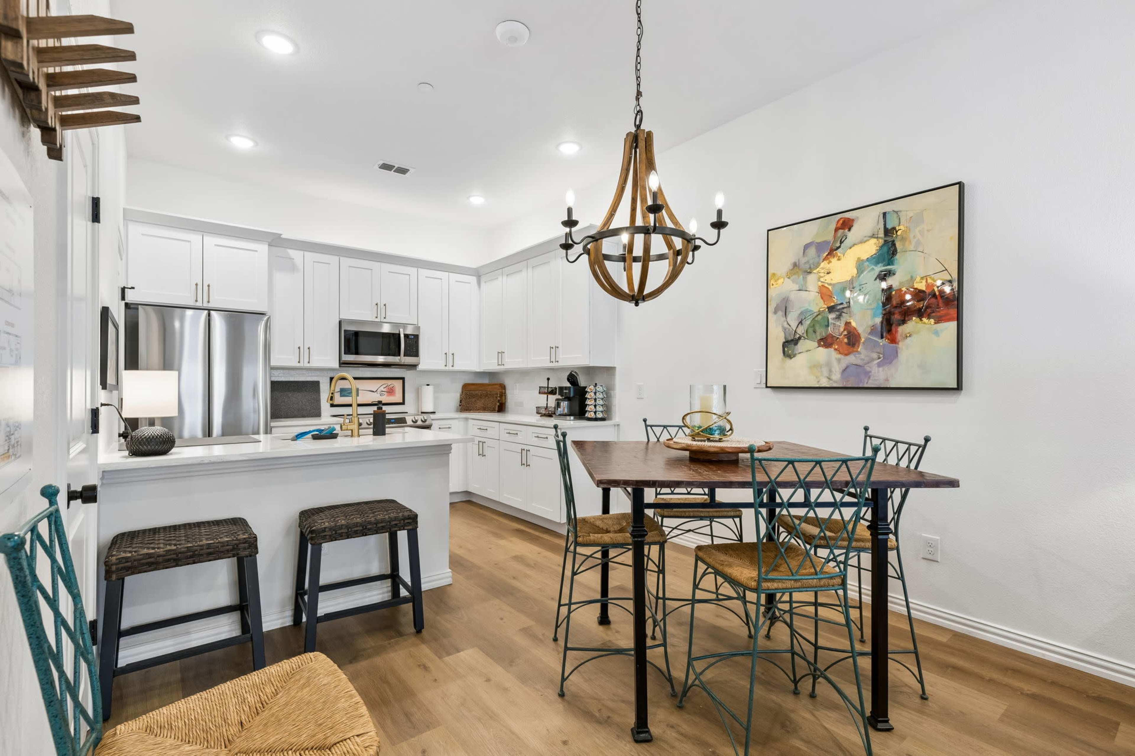 The image shows a modern kitchen and dining area featuring white cabinets, stainless steel appliances, a wooden dining table, and a chandelier.