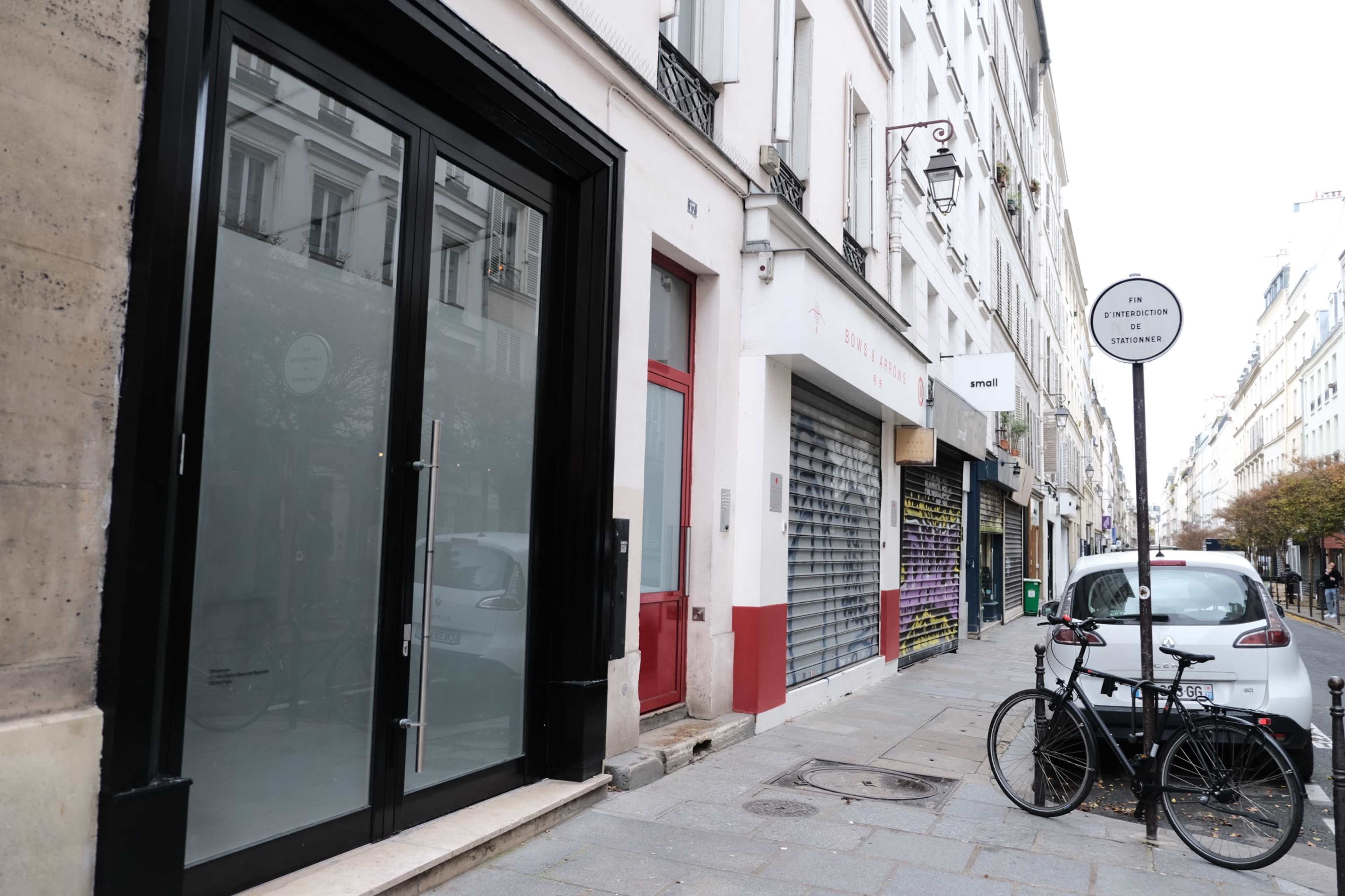 The image shows a street in Paris featuring a closed storefront with large glass windows, a parked bicycle, and a street sign indicating the location.