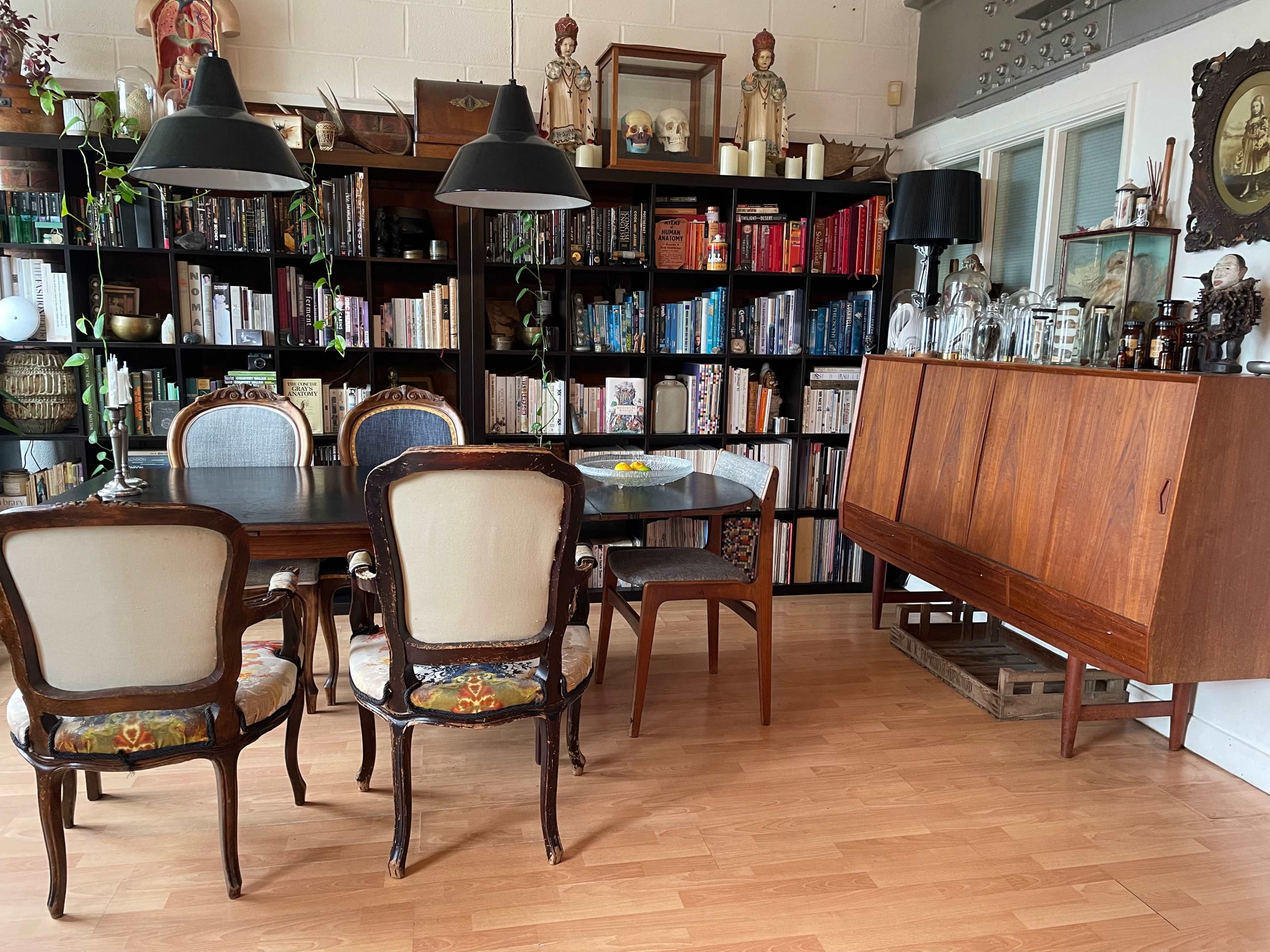 A dining area features a wooden table surrounded by chairs, with a bookshelf filled with various books and decorative items in the background.