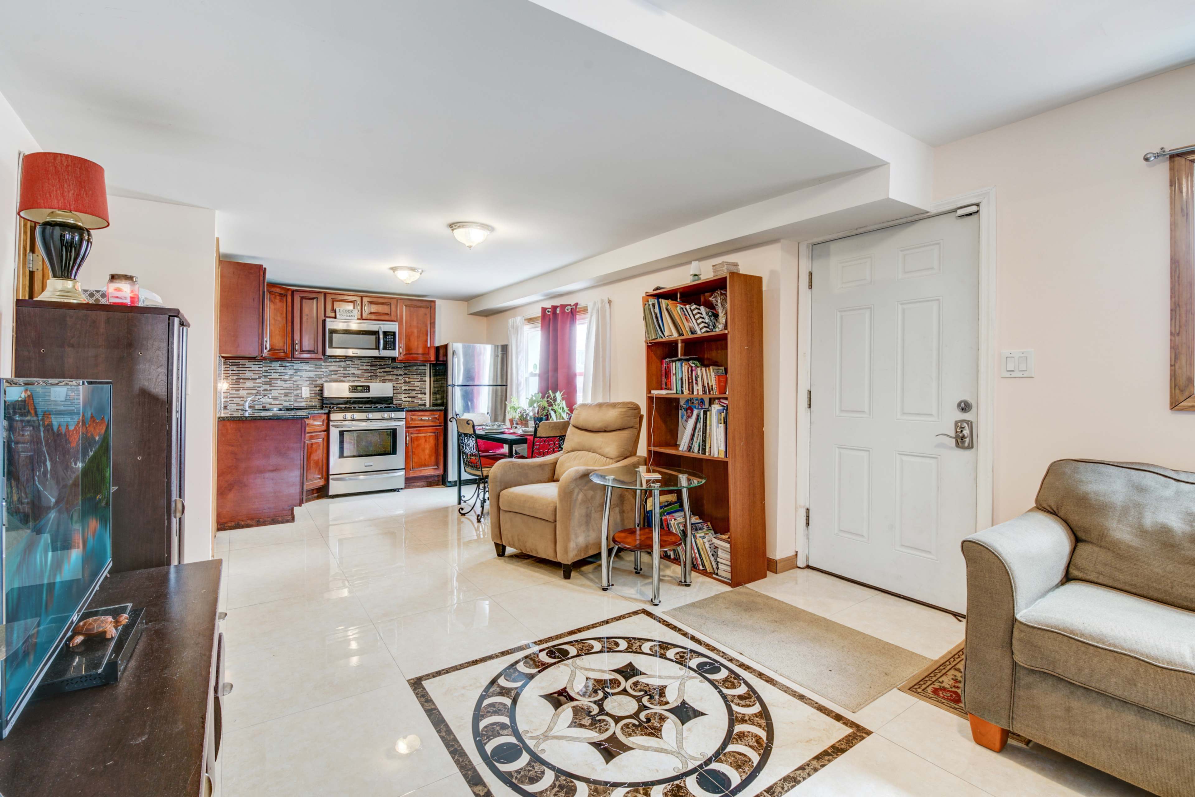 A compact living space features a kitchen with wooden cabinets, a plush armchair, a bookshelf, and a decorative area rug.