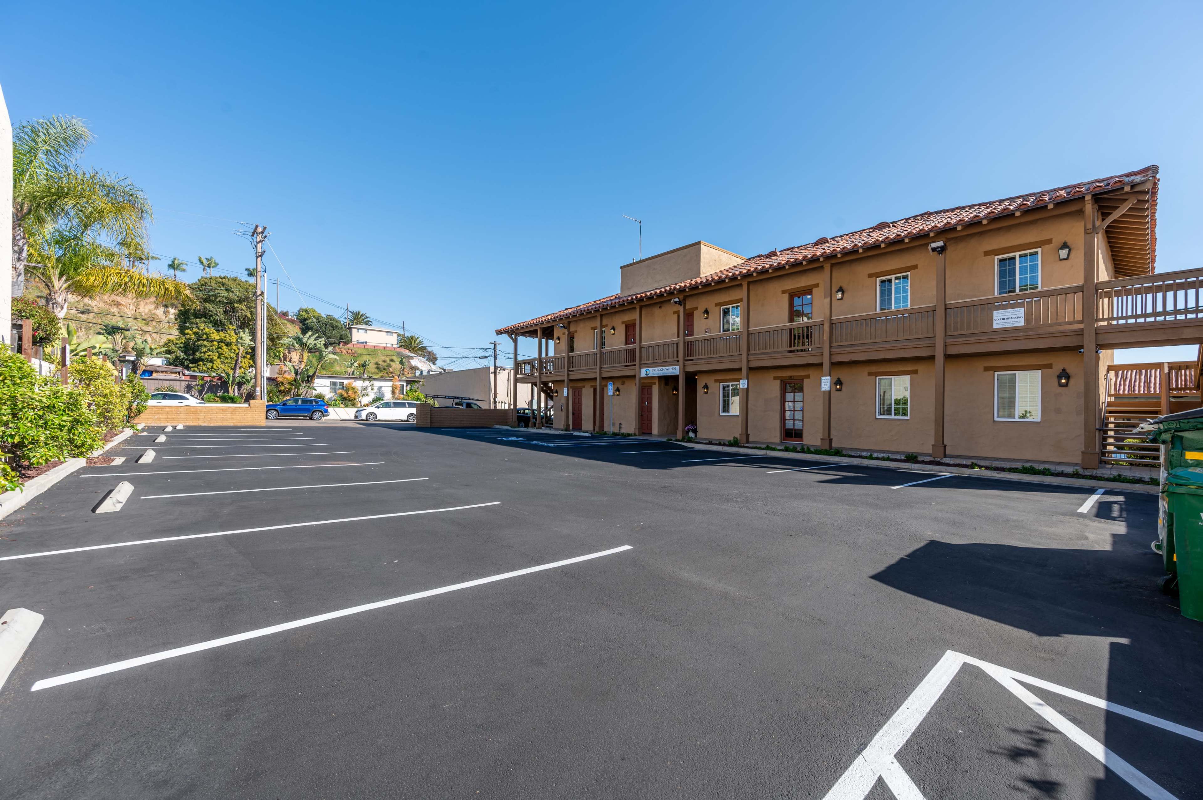 The image shows a vacant parking lot in front of a two-story motel with multiple rooms and a clear blue sky overhead.
