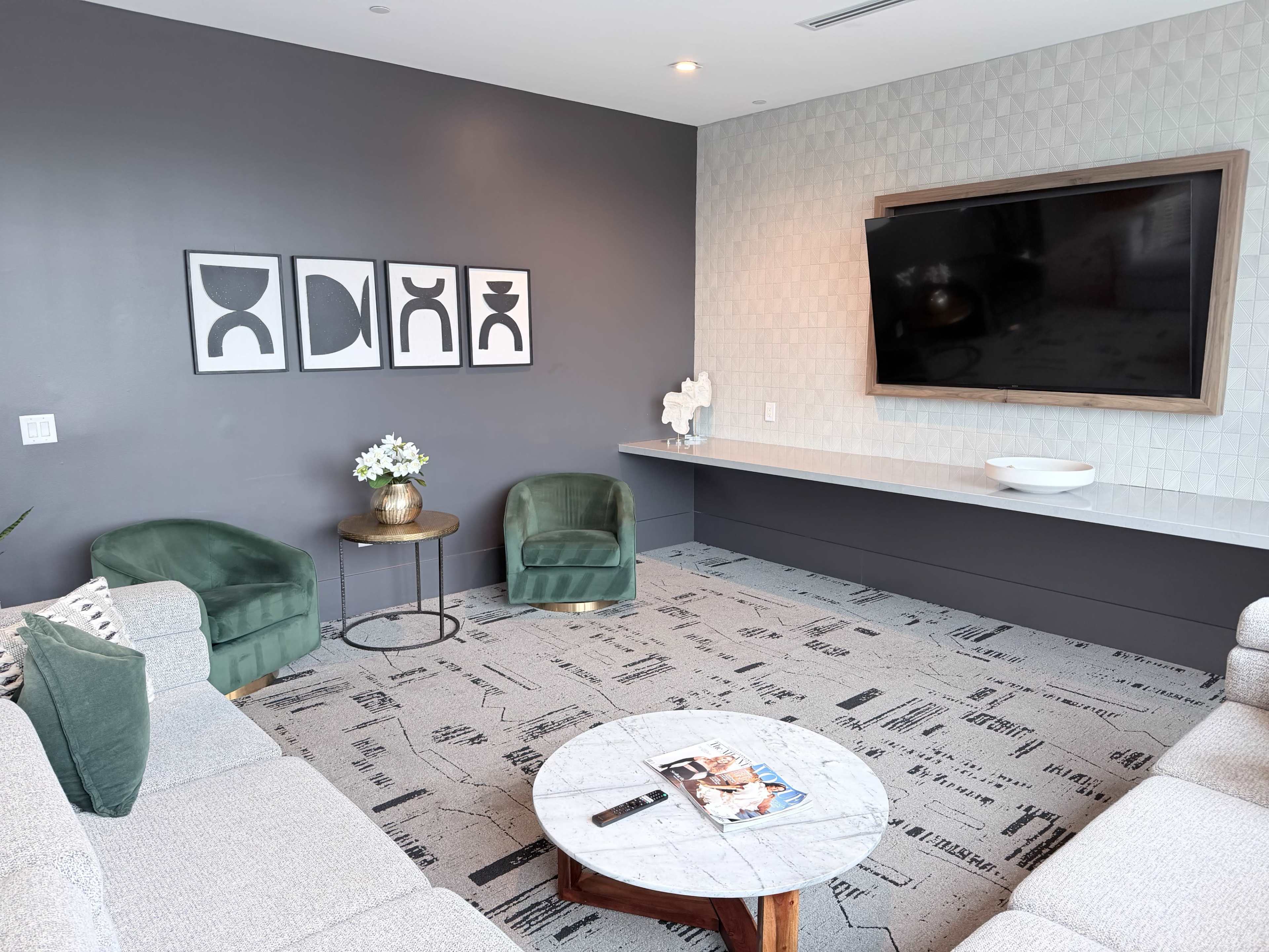 The image shows a modern living room with a grey feature wall, two green armchairs, a sofa, a round coffee table, and a large television mounted on the wall.
