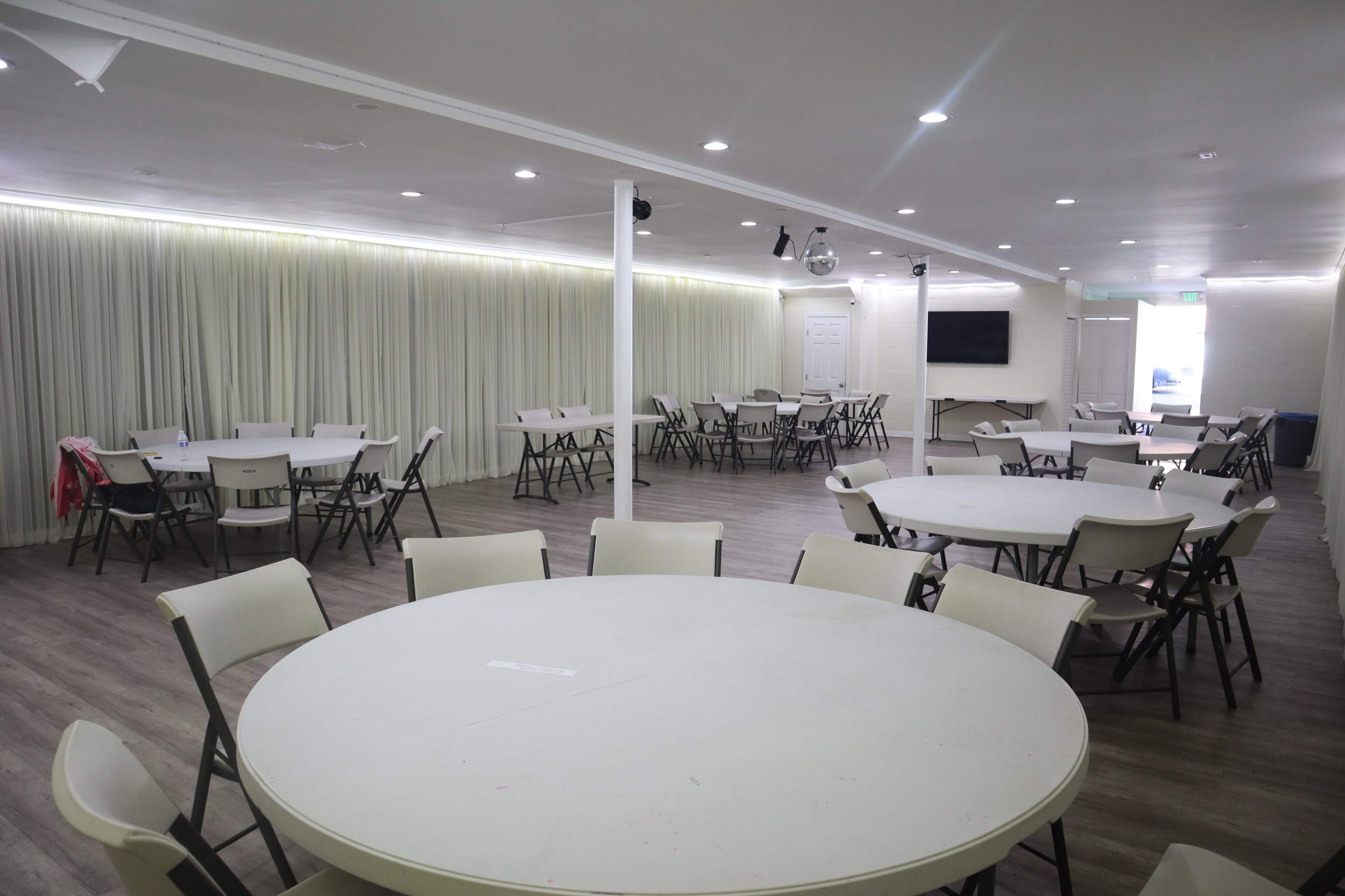 A large, empty event room features multiple round tables and chairs arranged neatly, with neutral-colored walls and curtains.