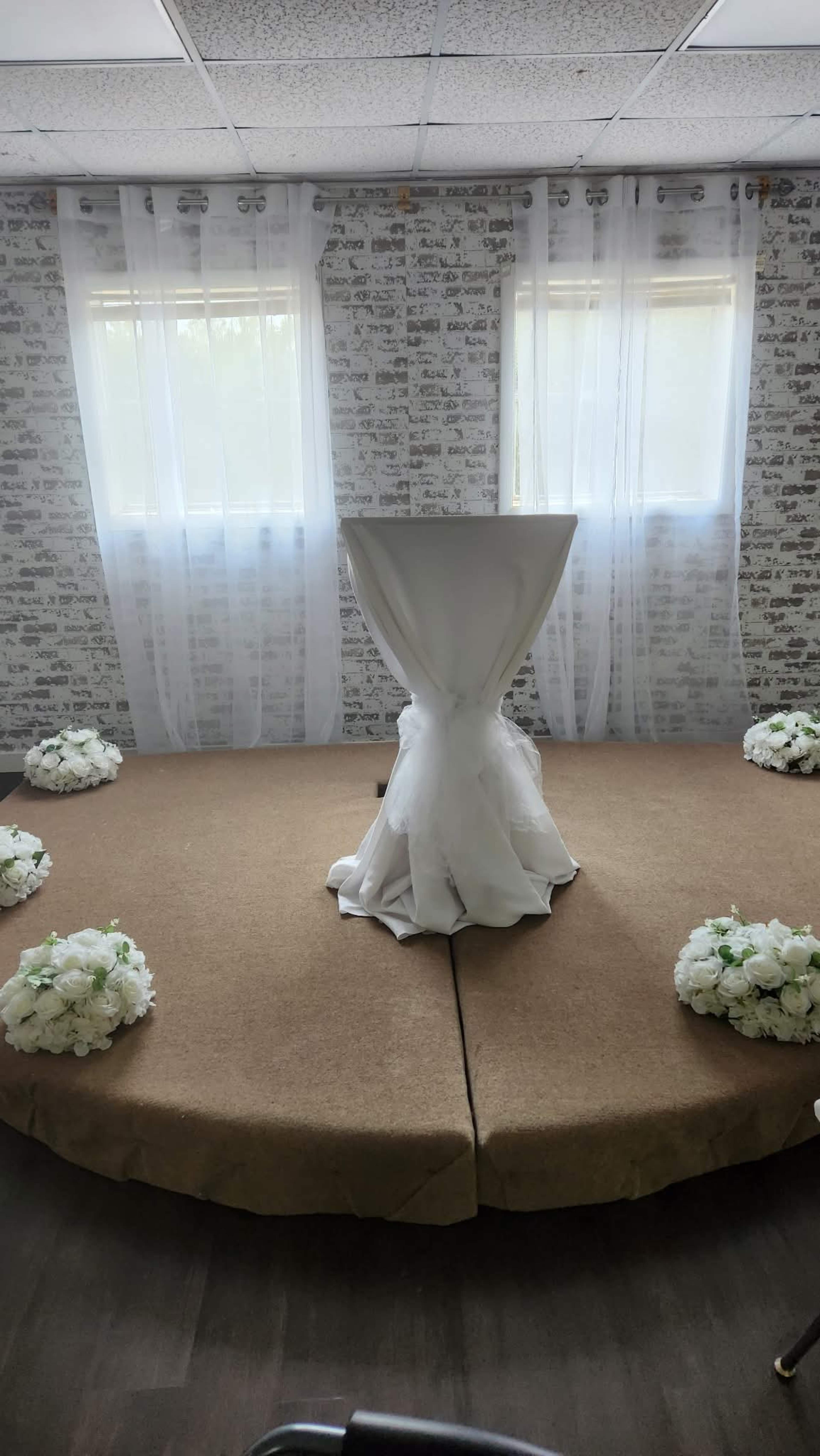 The image shows a setup for a wedding ceremony featuring a white table draped in fabric at the center, surrounded by floral arrangements on a beige carpet in a room with two windows covered by sheer curtains.
