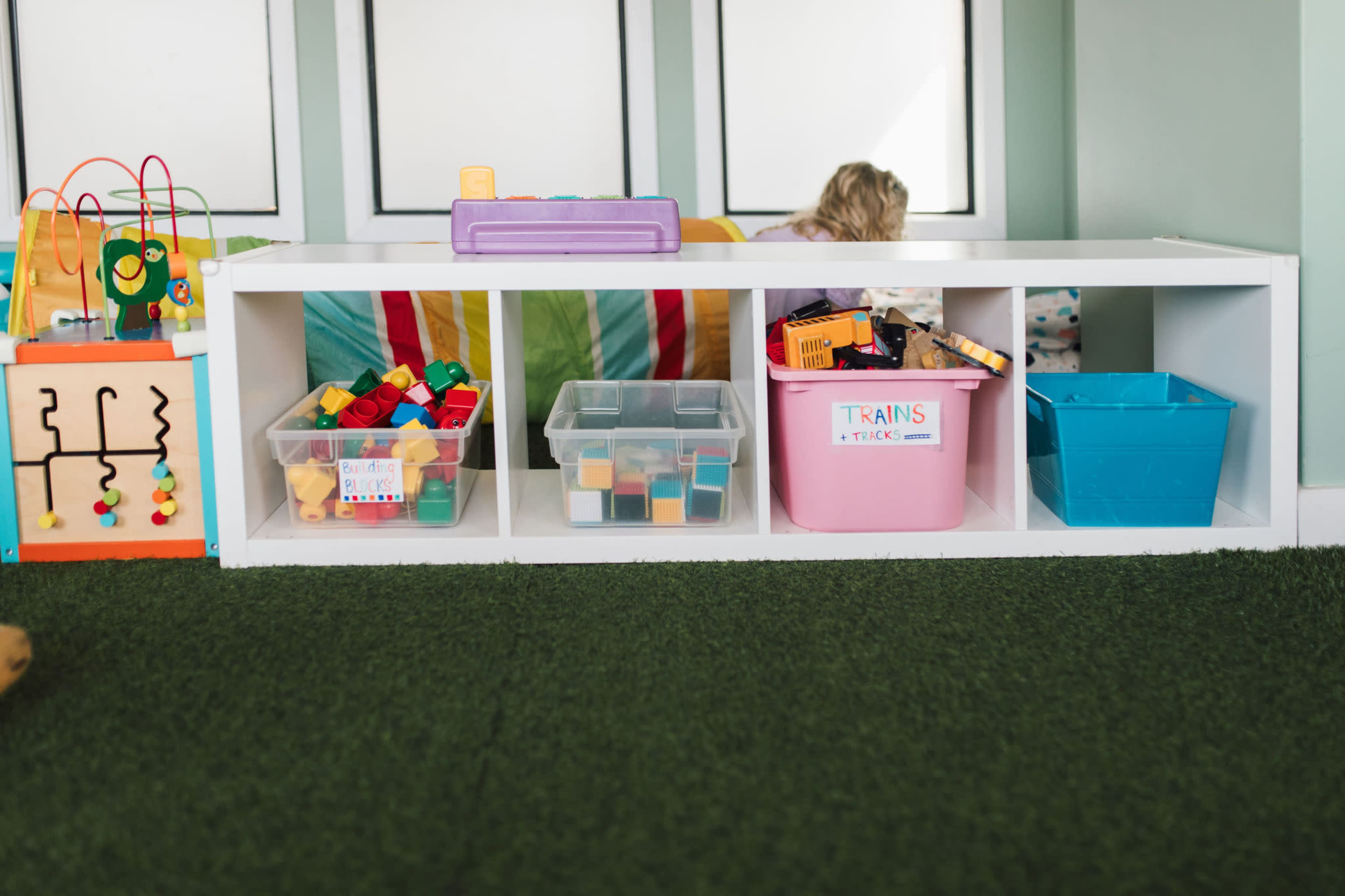 A play area features a white storage unit with bins of colorful toys and building blocks, set against a backdrop of green carpet and a colorful wall.