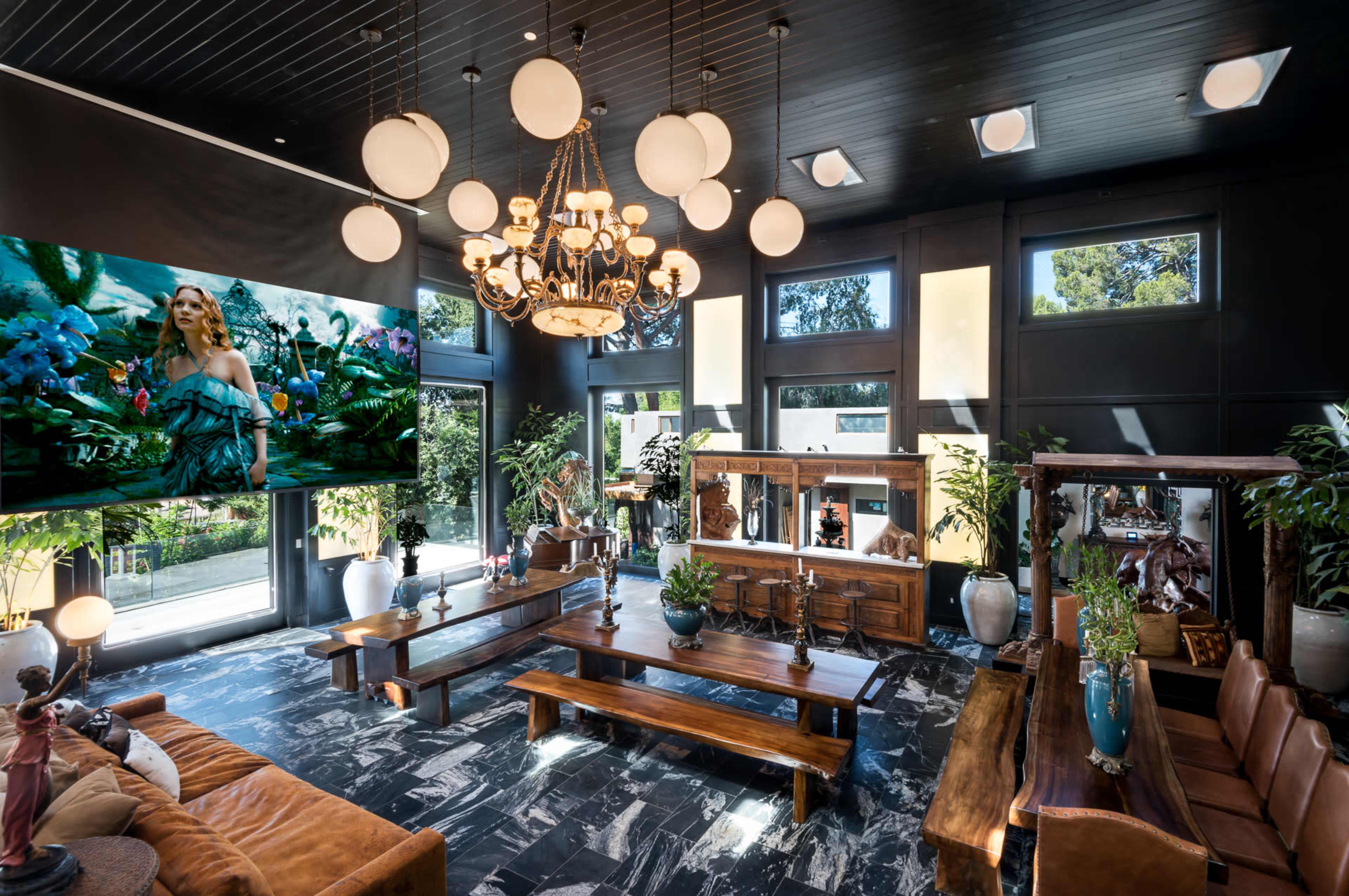 A spacious, modern living room featuring a large chandelier, a raised wooden table, and a big television screen displaying a colorful artwork among various indoor plants.