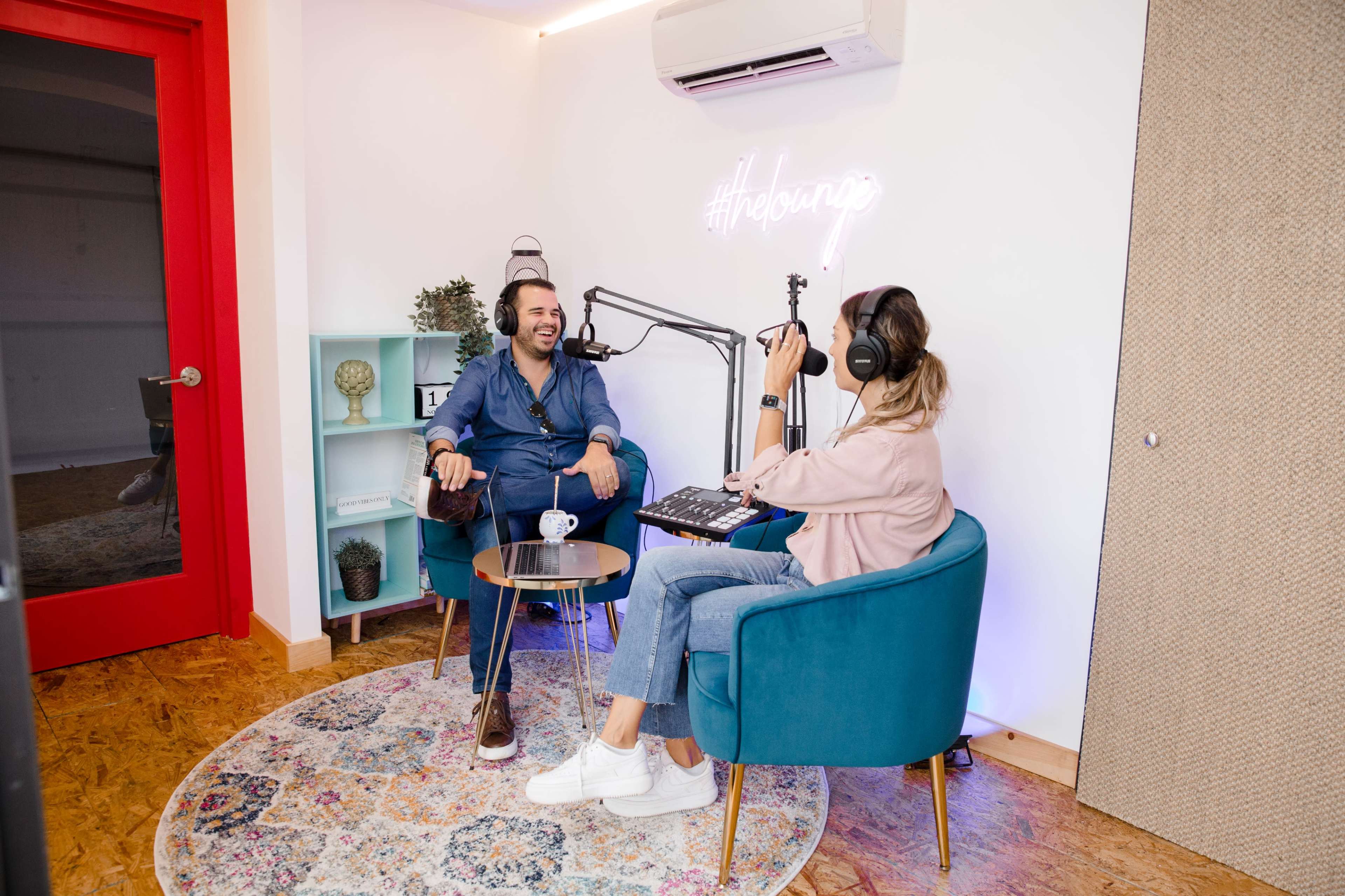 A man sits in a chair while a woman with headphones records a podcast, surrounded by colorful decor and a small table.