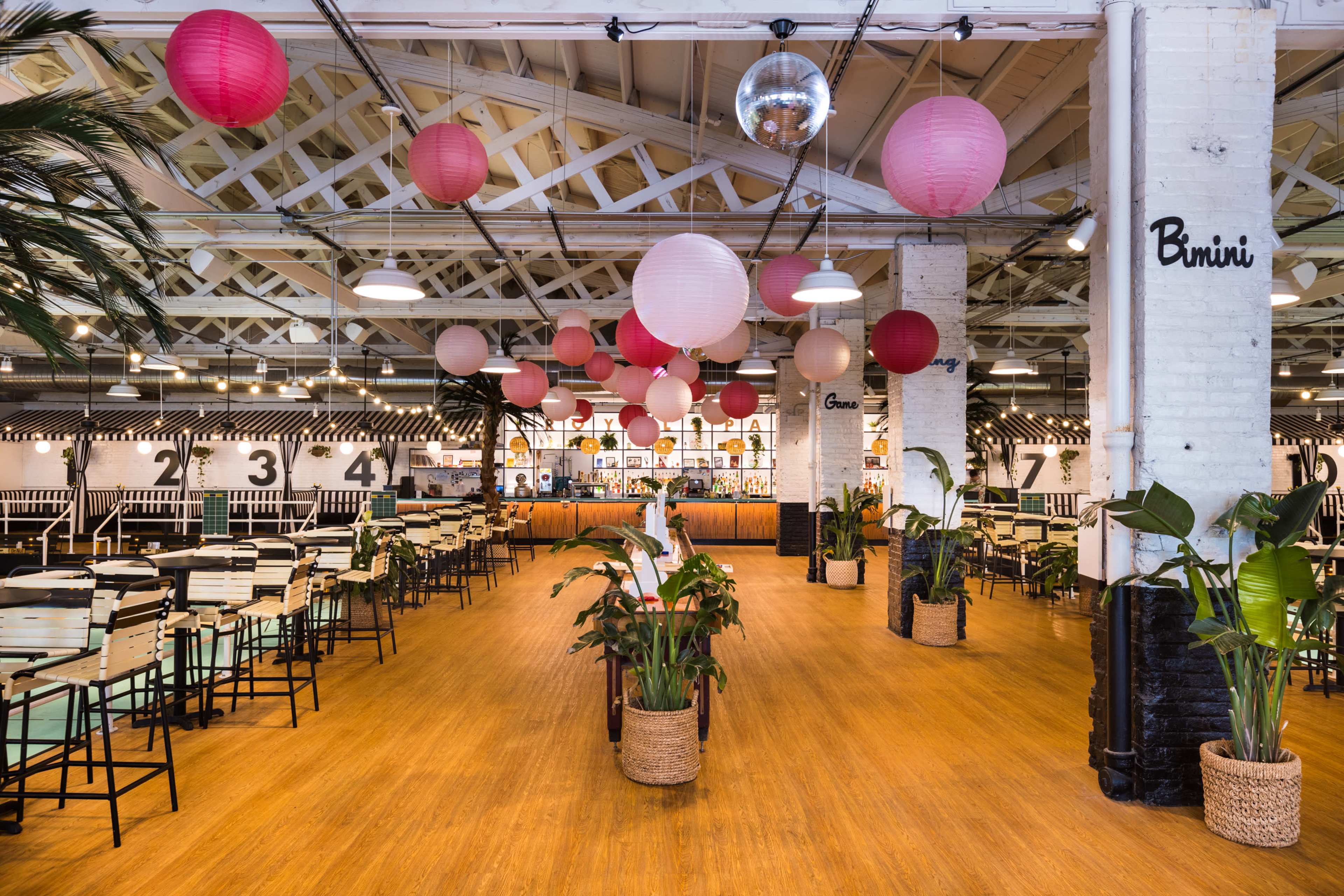 The image shows a spacious dining area with a bar at the back, decorated with hanging pink and red lanterns, potted plants, and wooden furniture.