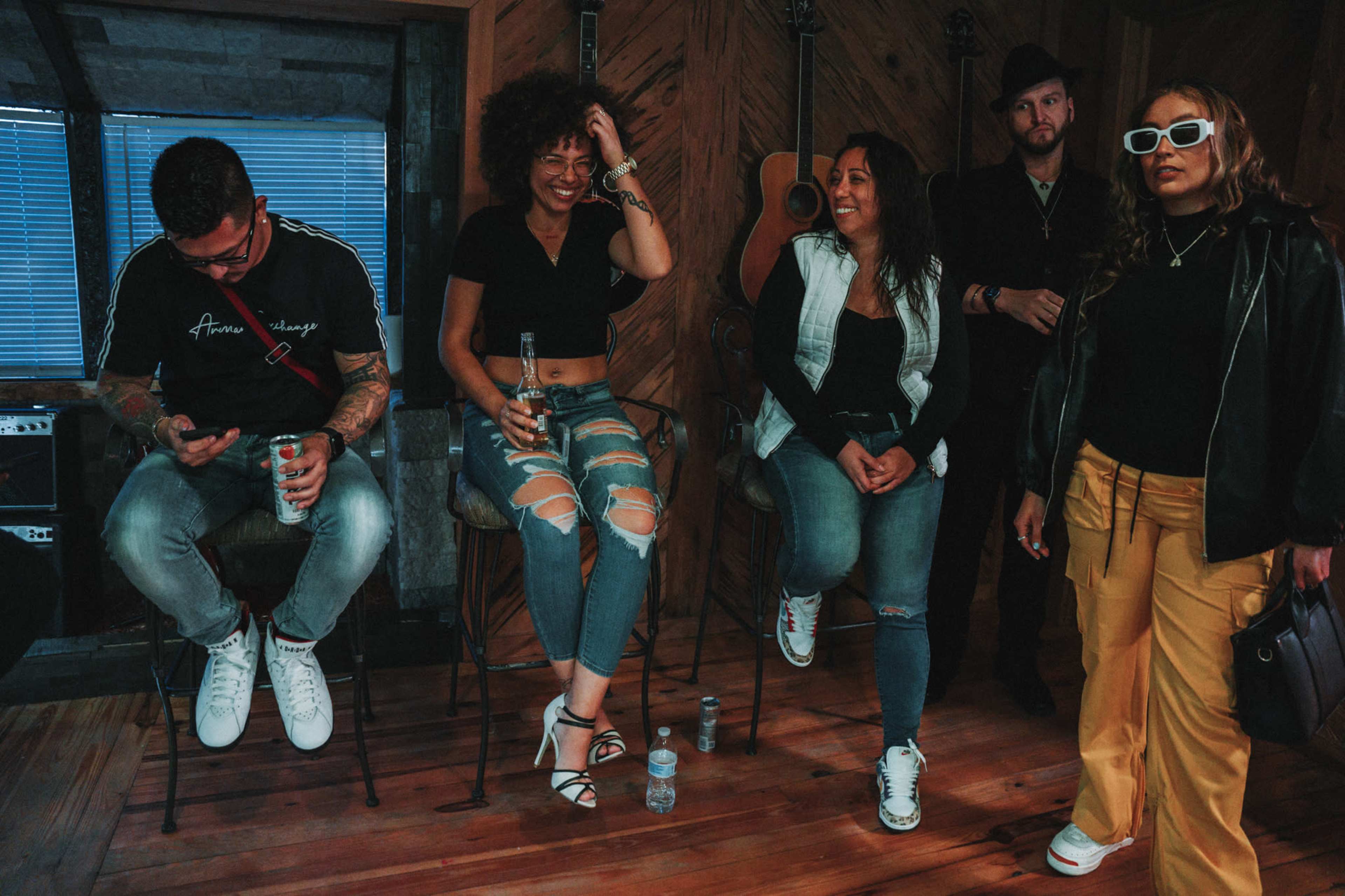 A group of five people sits in a wooden room with musical instruments, discussing and laughing while enjoying drinks.