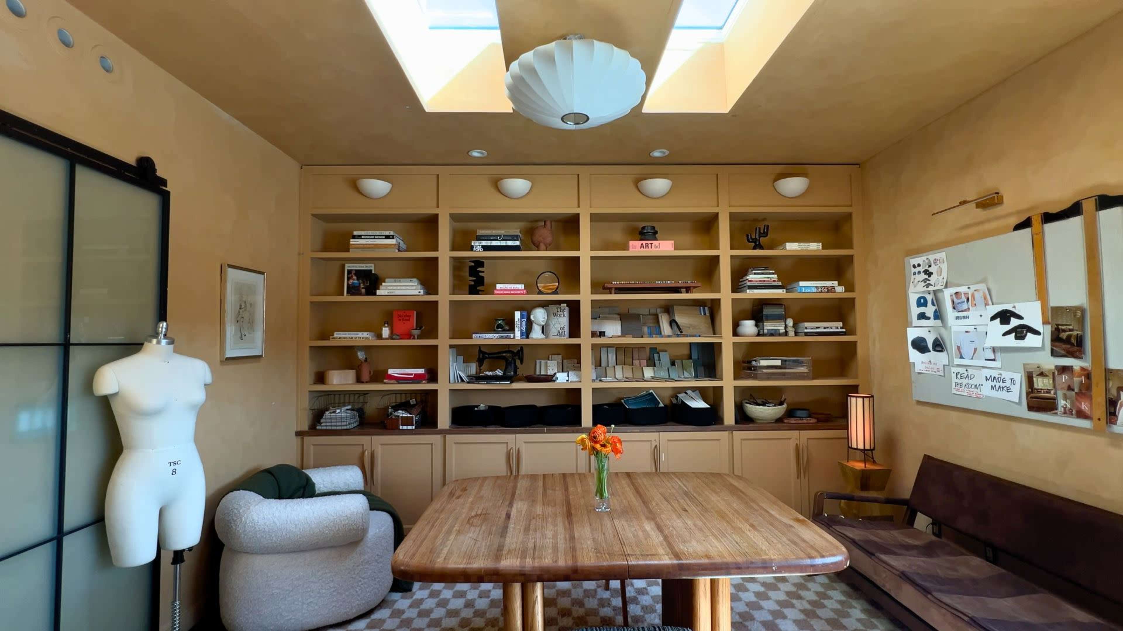 The image shows a well-organized room with a wooden table at its center, surrounded by shelves filled with various items and a mannequin in the corner.