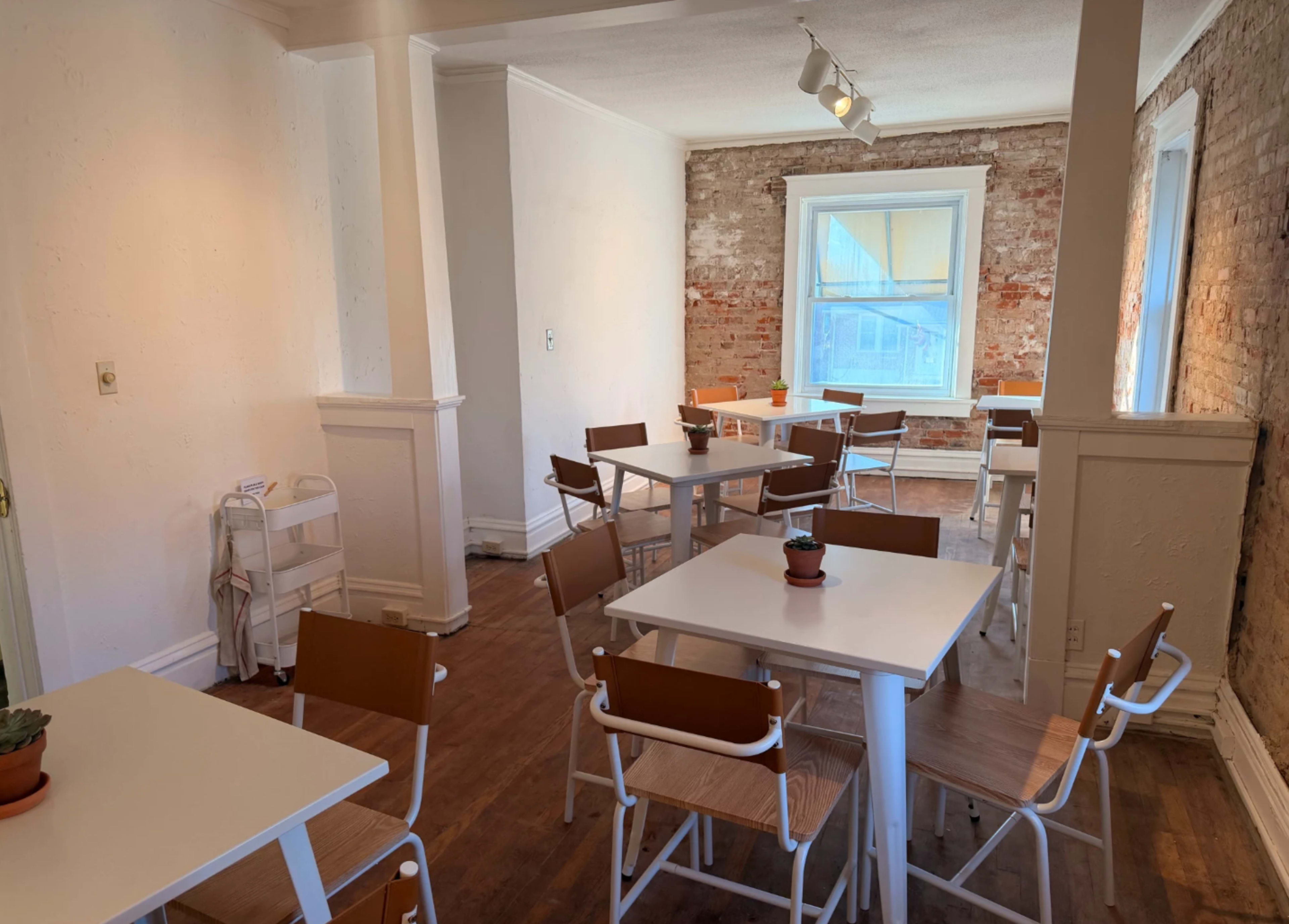The image shows a lightly furnished dining area with several white tables and wooden chairs arranged near a large window that allows natural light to enter, and exposed brick walls.