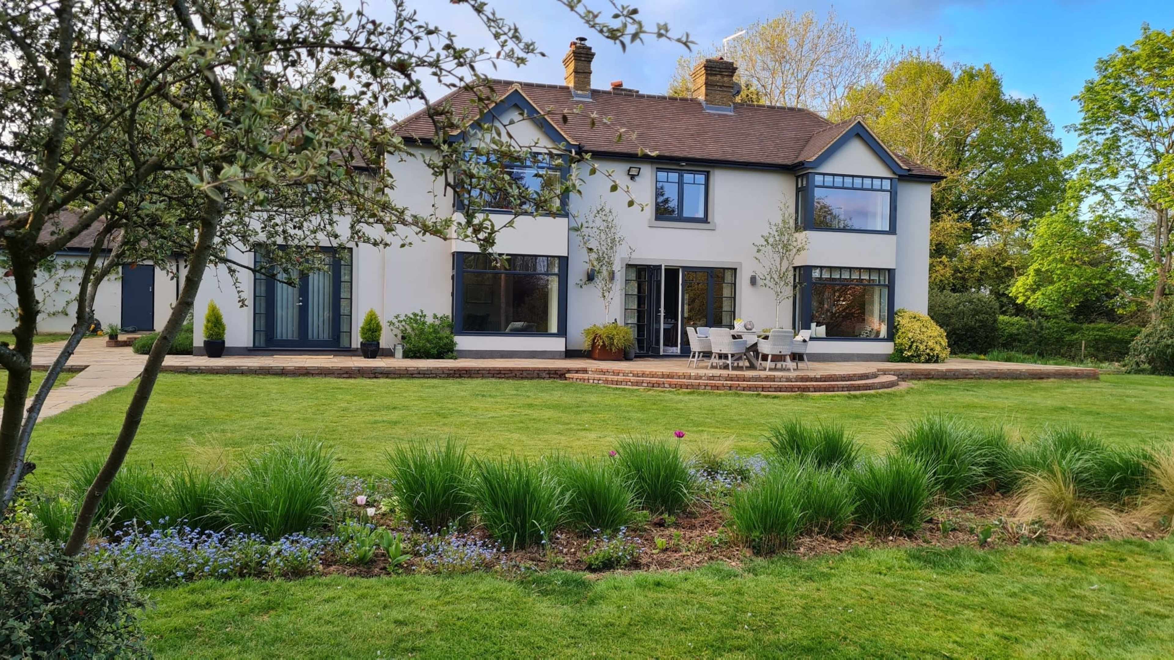 The image shows a two-story white house with a landscaped garden featuring a patio and seating area.