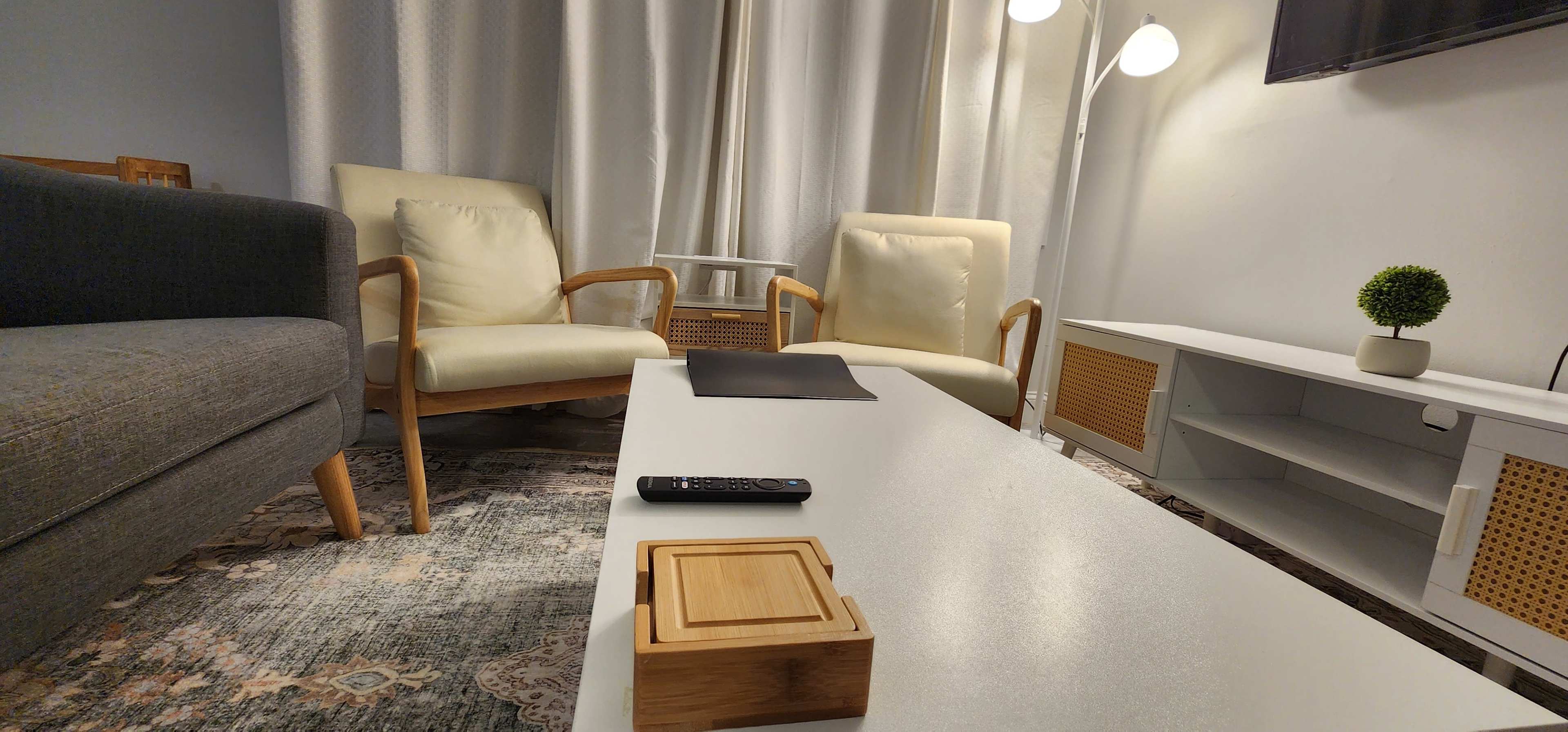 A living room with two cream-colored armchairs, a gray sofa, a coffee table with a remote control and a decorative wooden box, and a floor lamp beside a window.