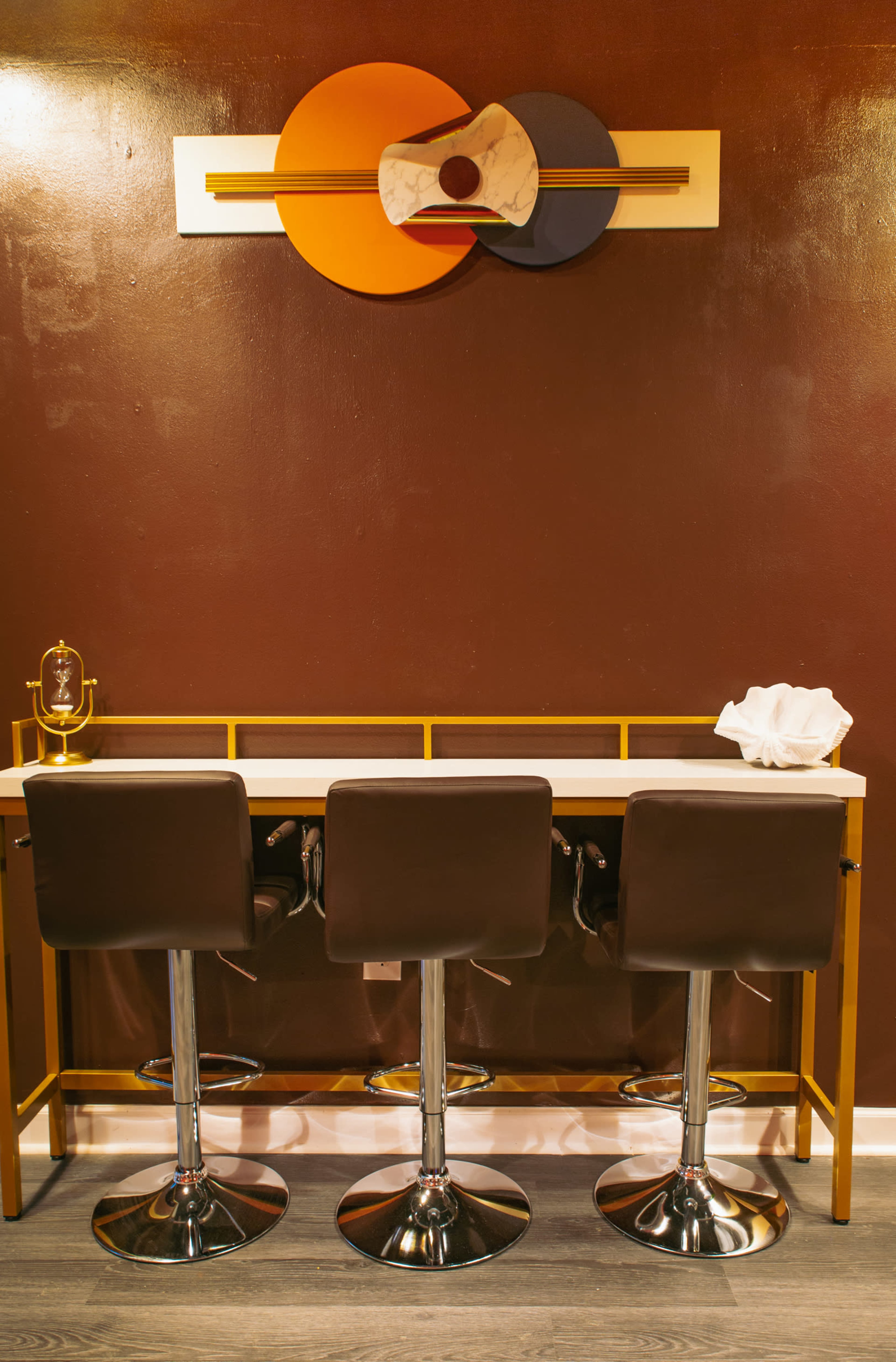 A minimalist bar area features three high stools in front of a simple counter, complemented by a geometric wall art piece above.