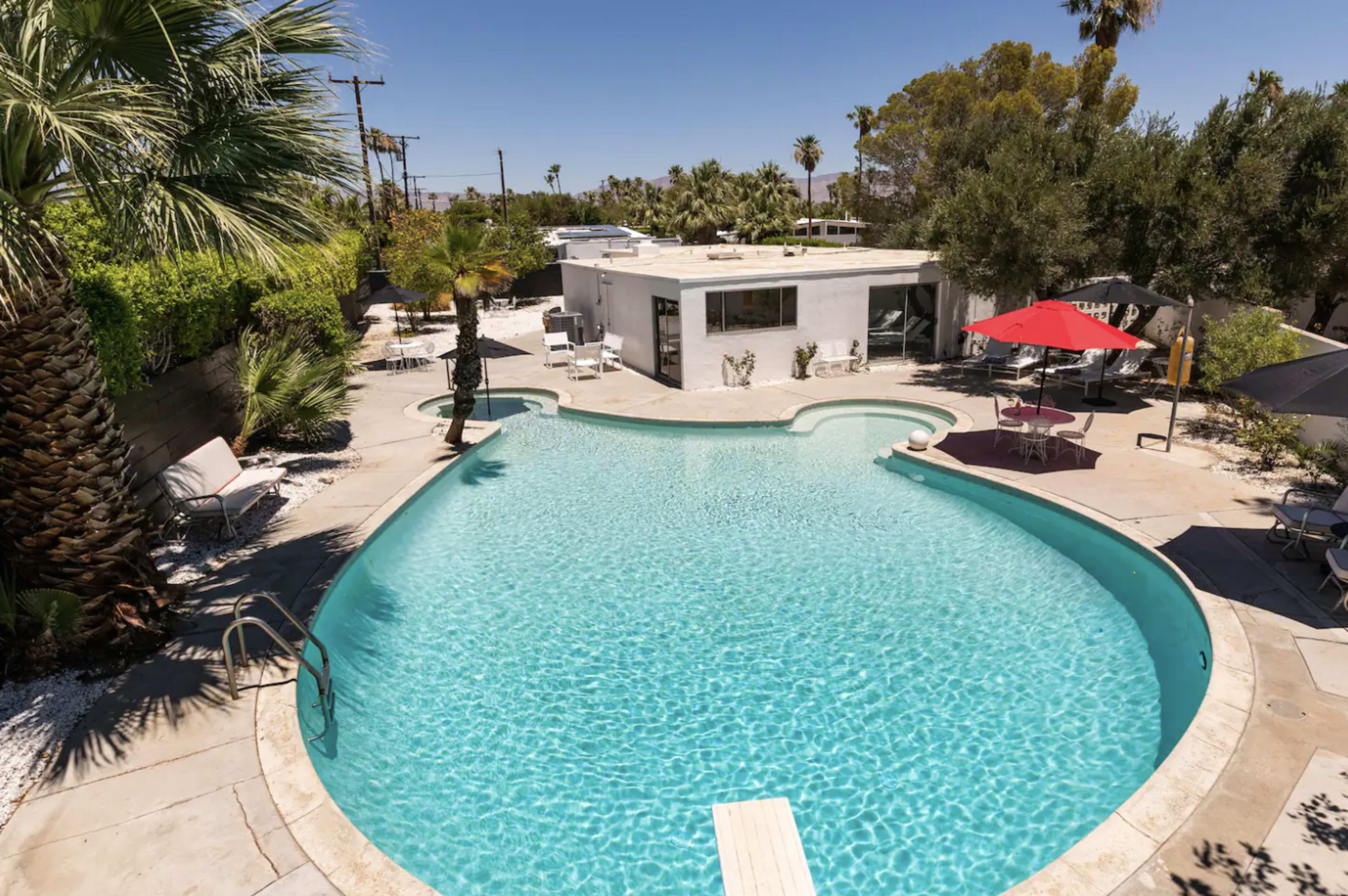 A curved swimming pool surrounded by palm trees and lounge chairs, with a bright red umbrella near a patio area.