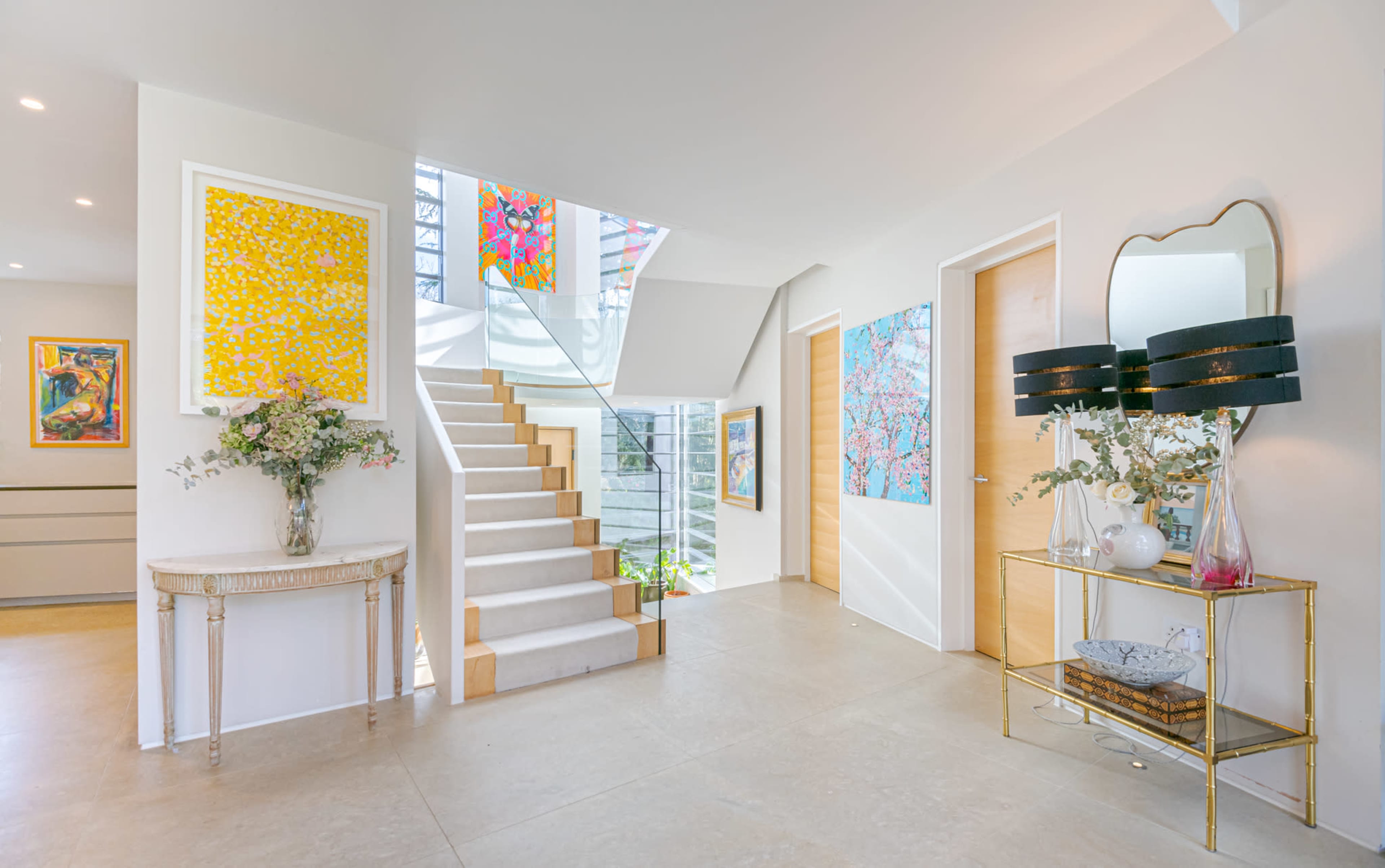 The image shows a modern entryway with a staircase, adorned with colorful artwork and a table displaying floral arrangements and decorative items.