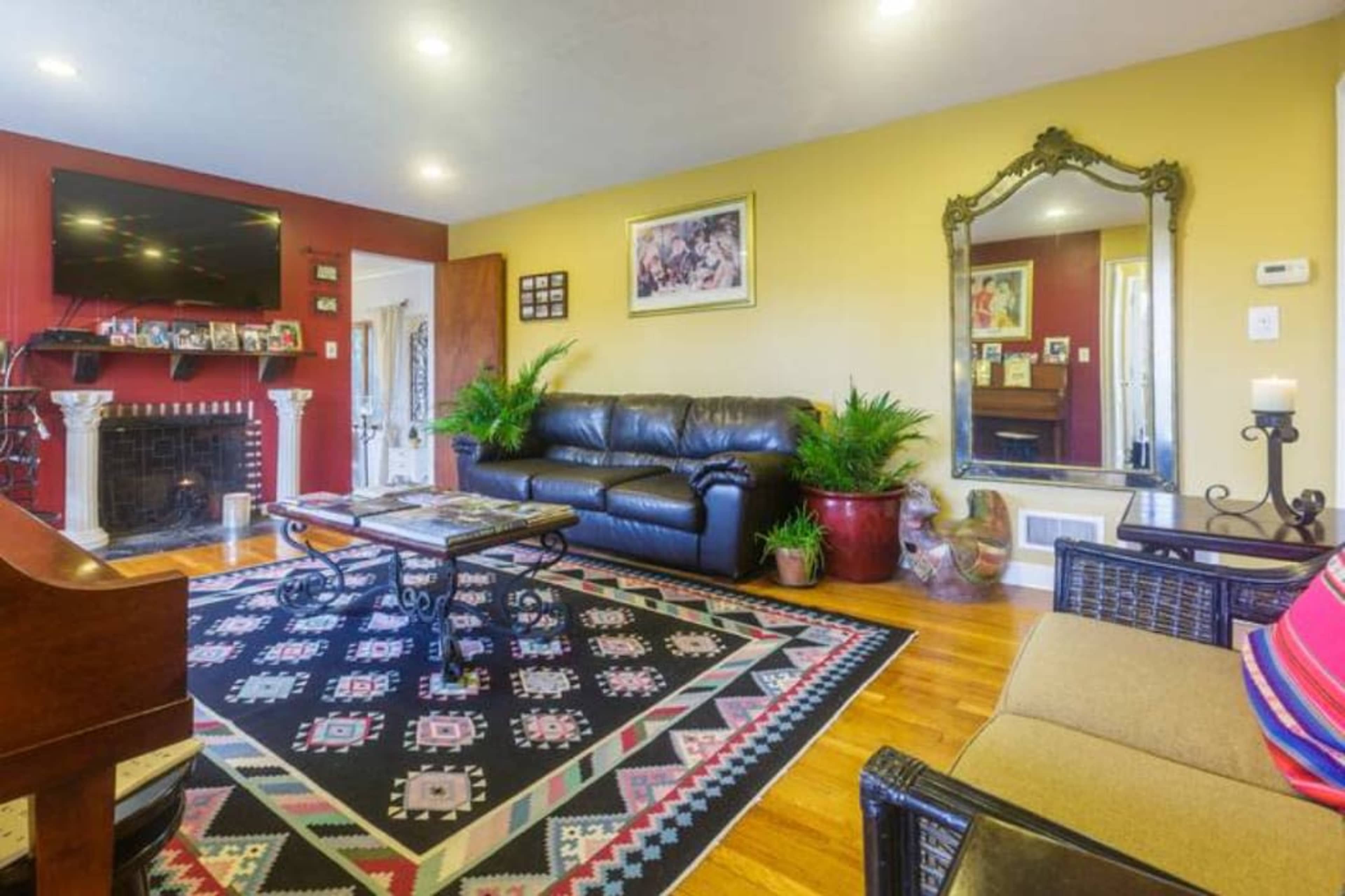 The living room features a black leather sofa, a patterned area rug, a wooden coffee table, and a large mirror on the wall, with plants and decorative elements throughout.