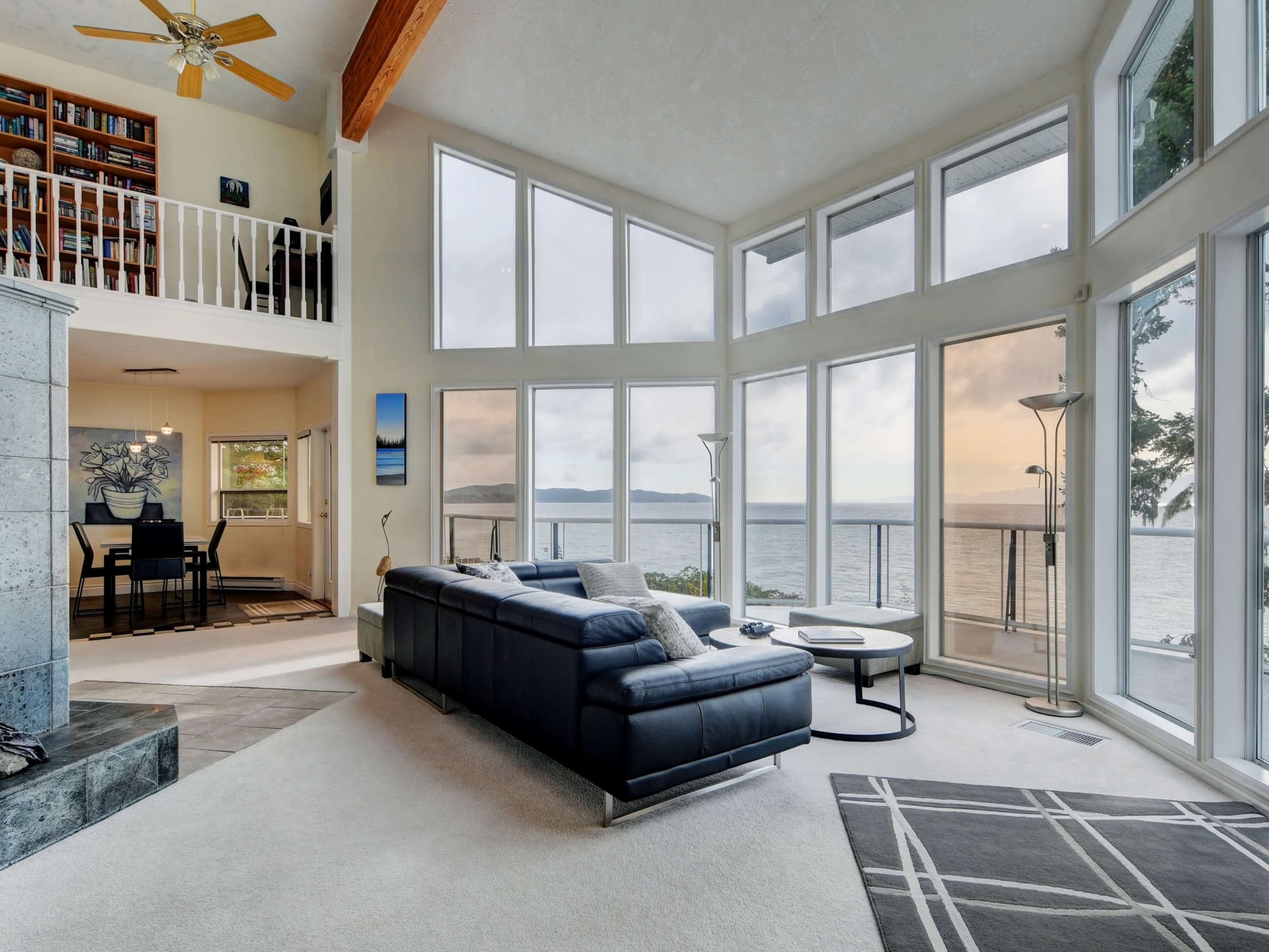A modern living room features large windows overlooking the ocean, with a sectional sofa, coffee table, and a bookshelf in the background.