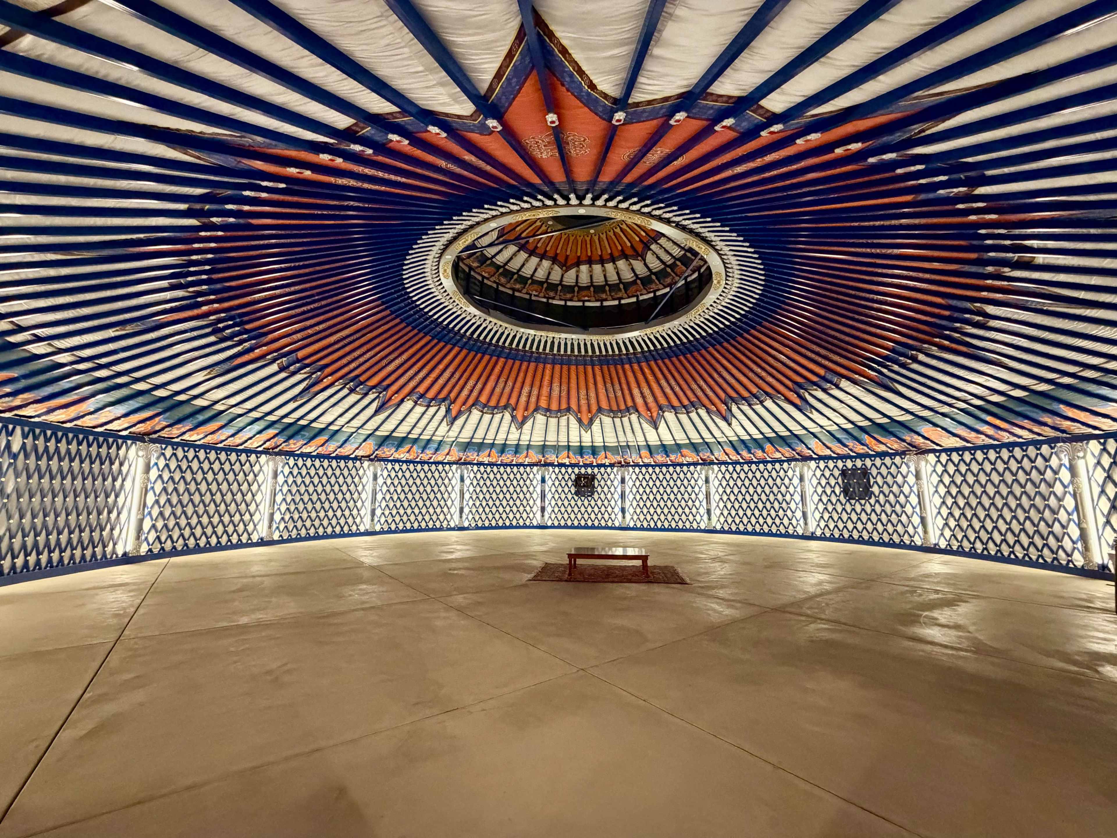 The interior of a yurt features a circular ceiling adorned with a colorful geometric pattern and light fixtures, surrounded by white walls decorated with blue and orange accents.