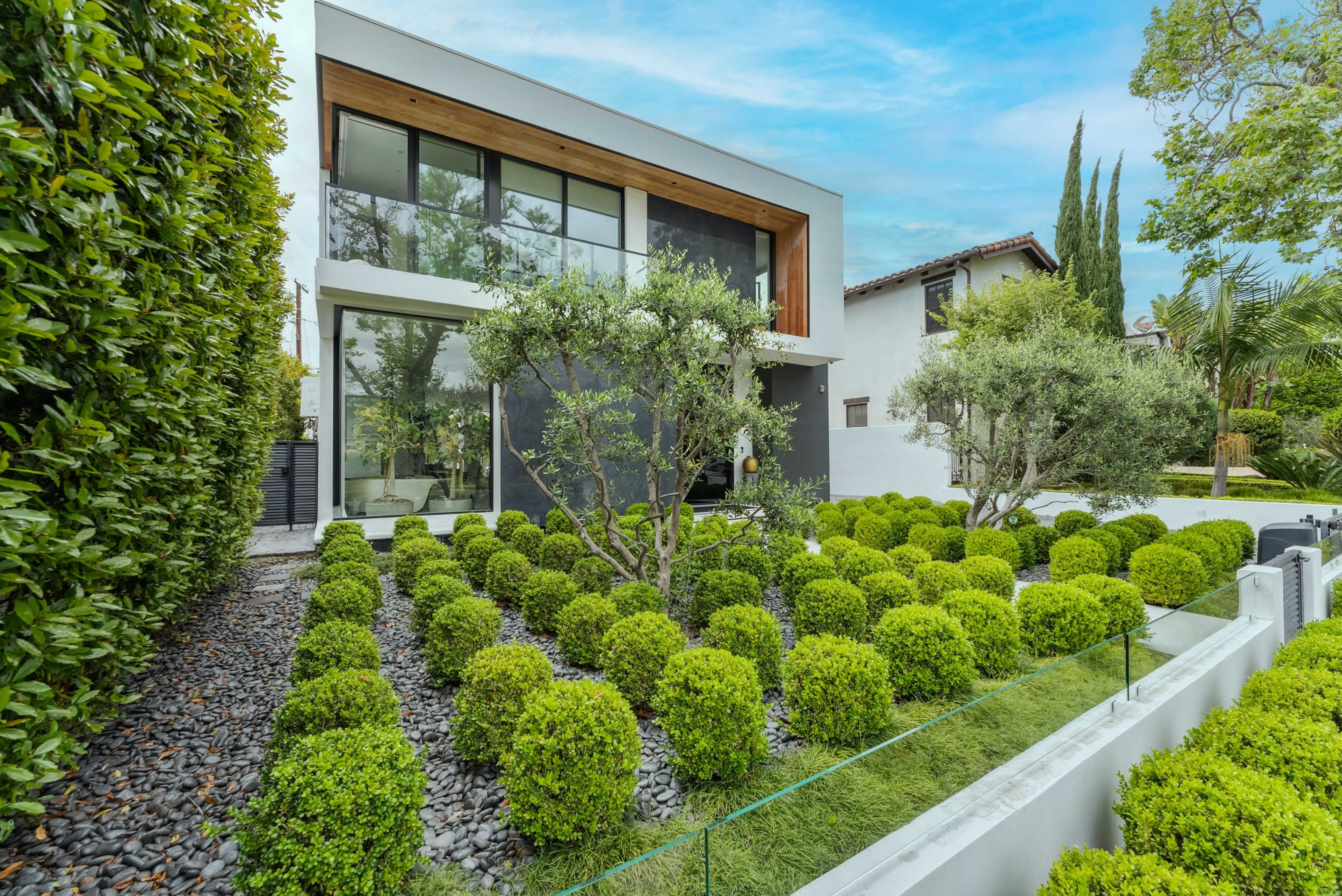 A modern house with large windows is surrounded by neatly manicured green hedges and stone pathways.