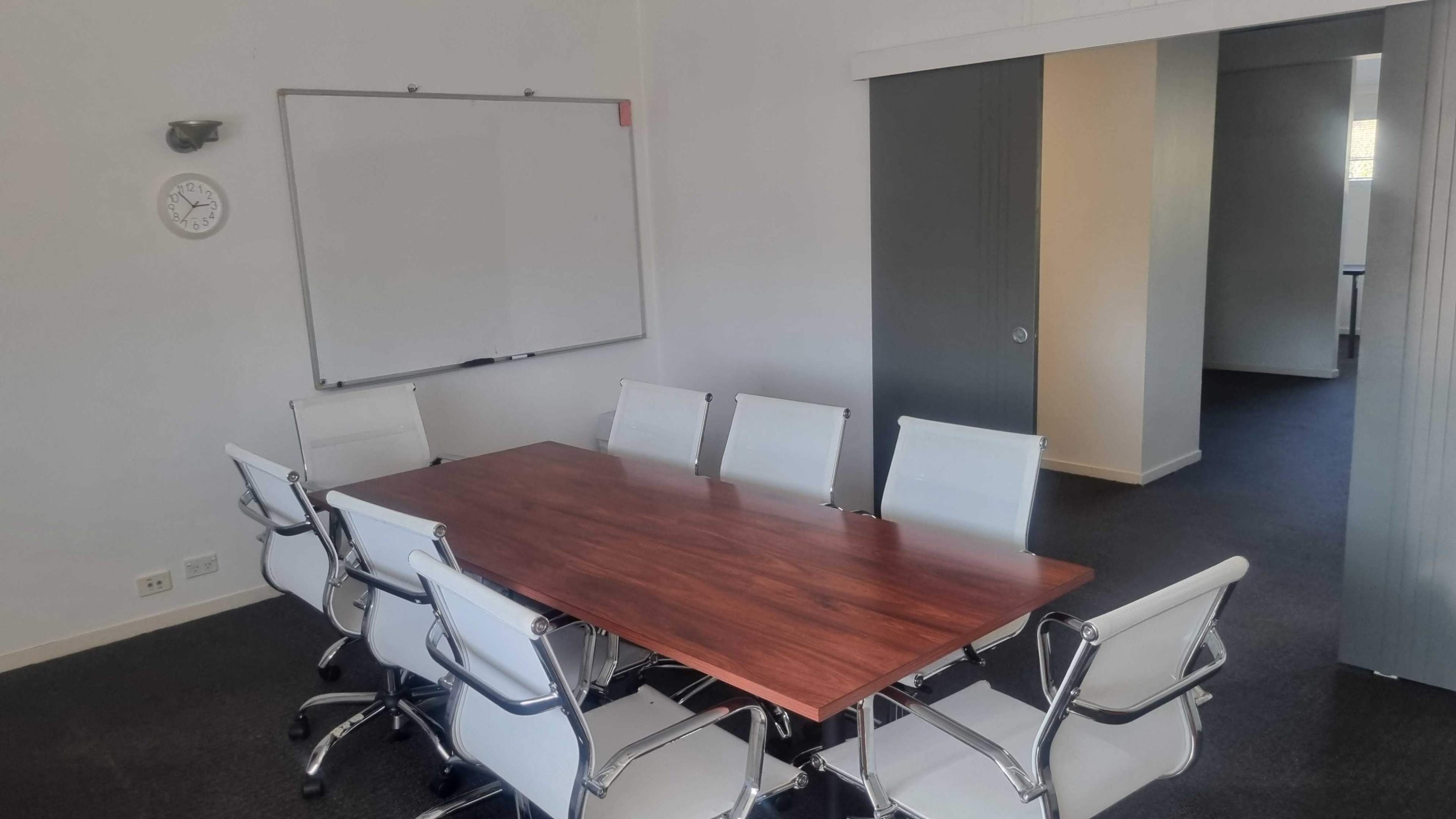 The image shows a conference room with a large wooden table surrounded by eight white chairs, a whiteboard on the wall, and a clock.