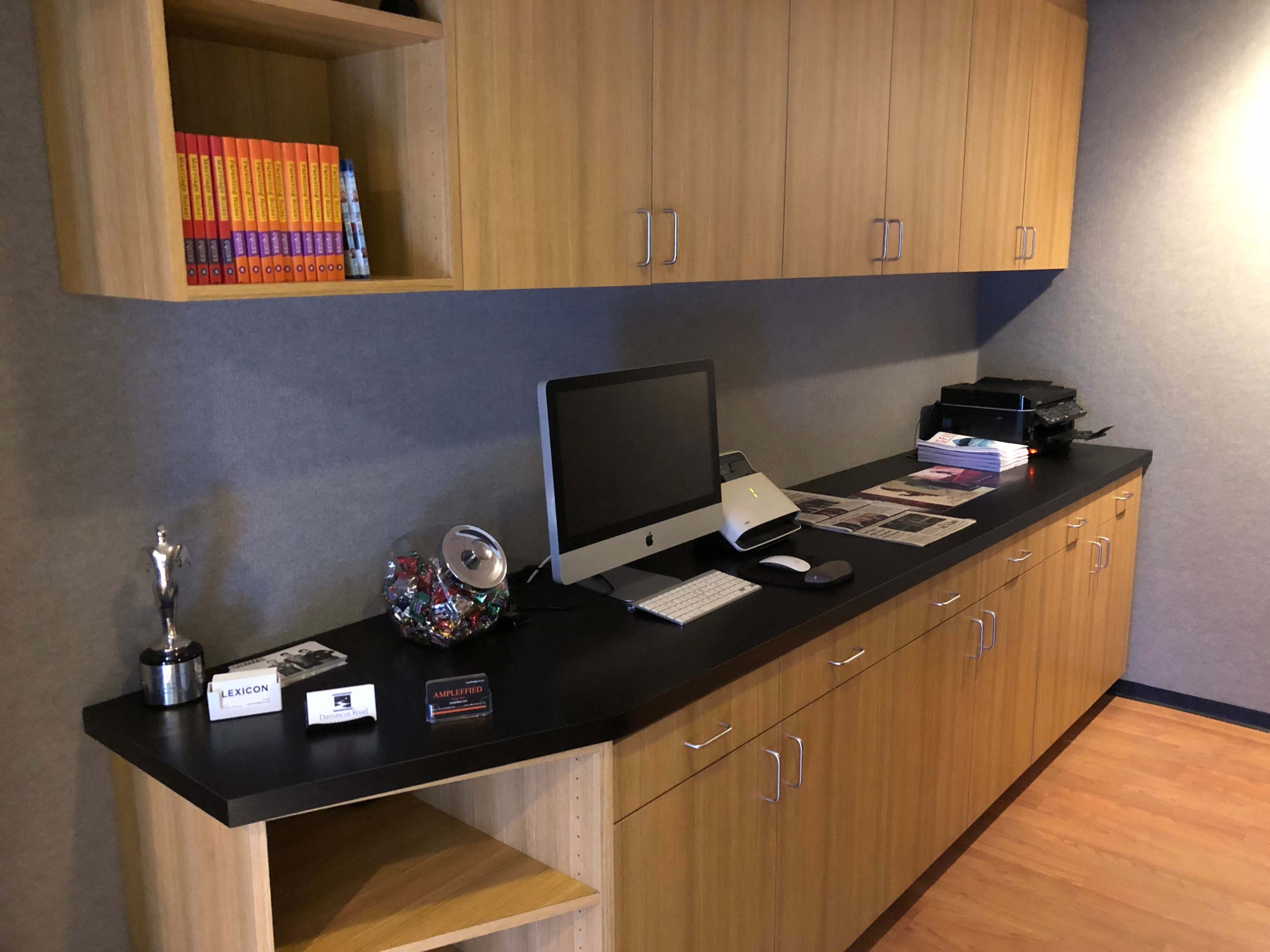 A wooden office space featuring a black countertop with a computer, printer, and various books and awards displayed on shelves.