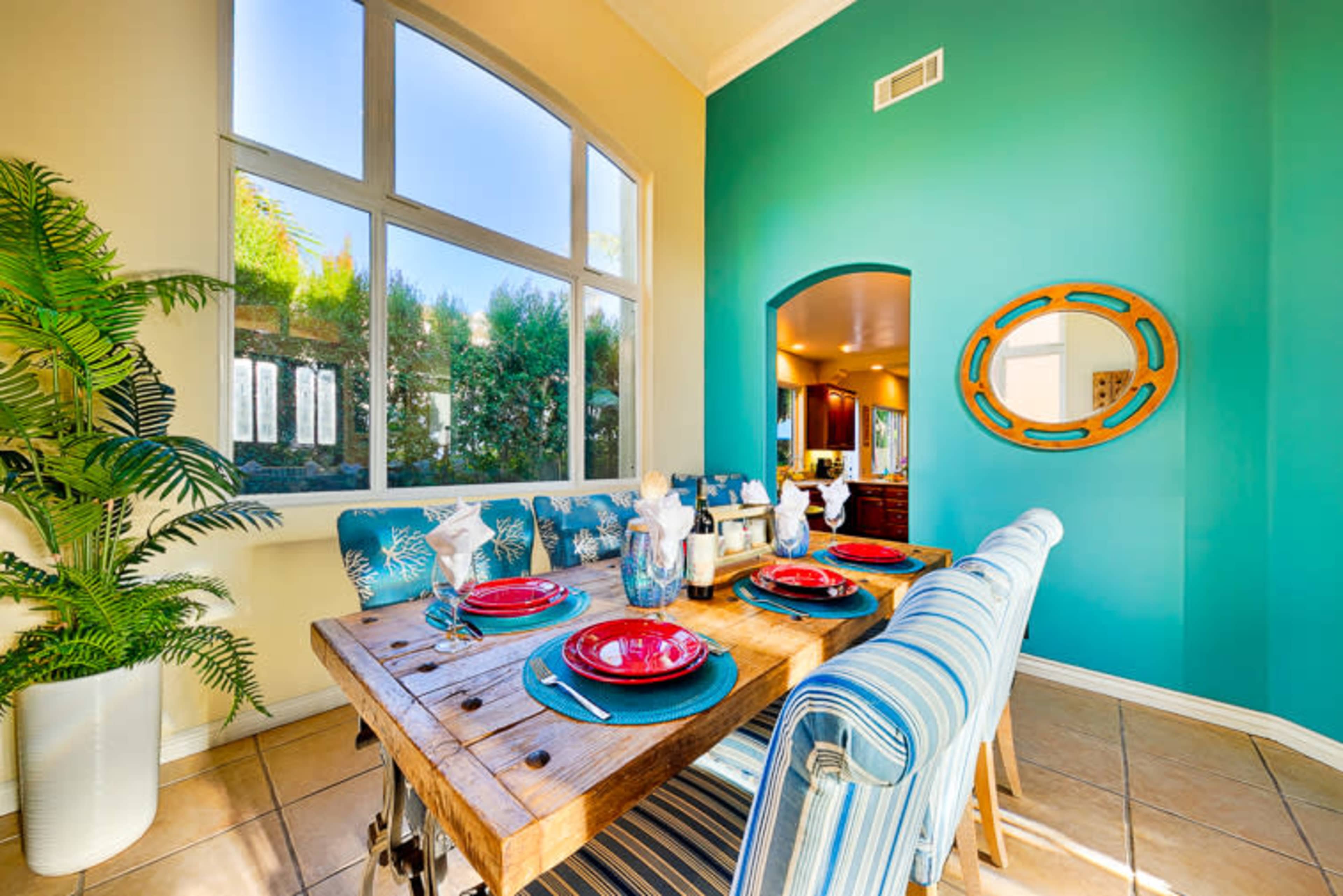 A dining area features a wooden table set with red plates and striped blue chairs, adjacent to large windows and a vibrant teal wall.