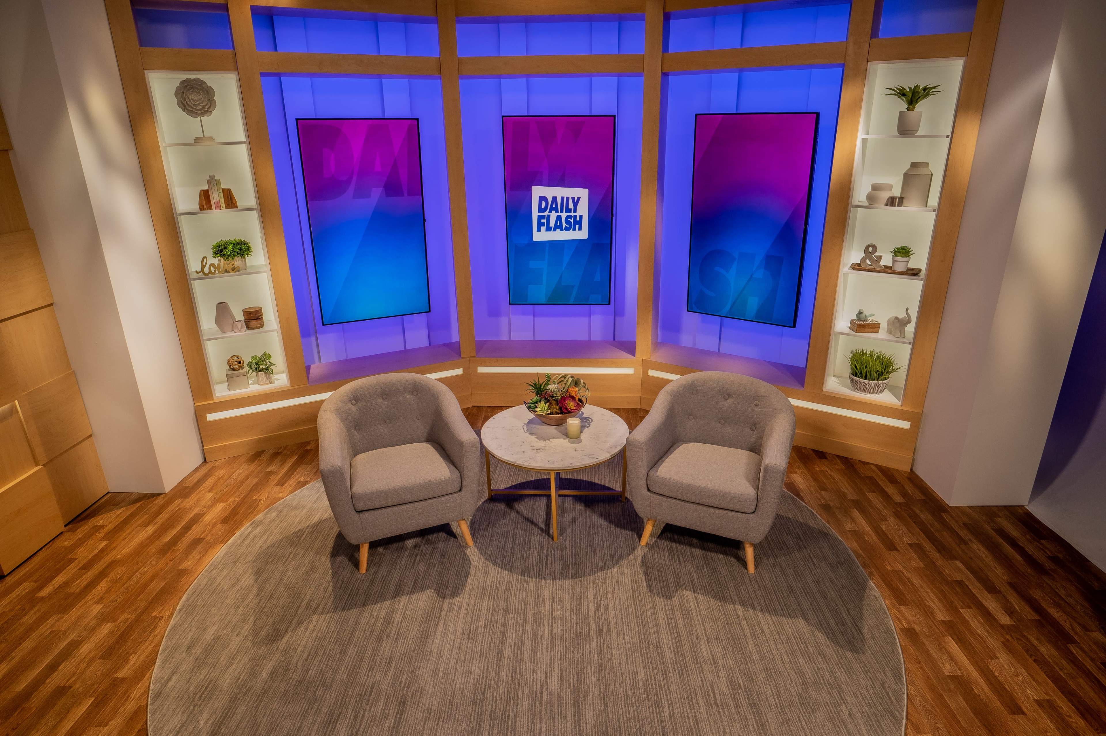 The image shows a television studio setup featuring two gray armchairs positioned around a circular coffee table, with a backdrop displaying the words "Daily Flash" against blue and purple screens.