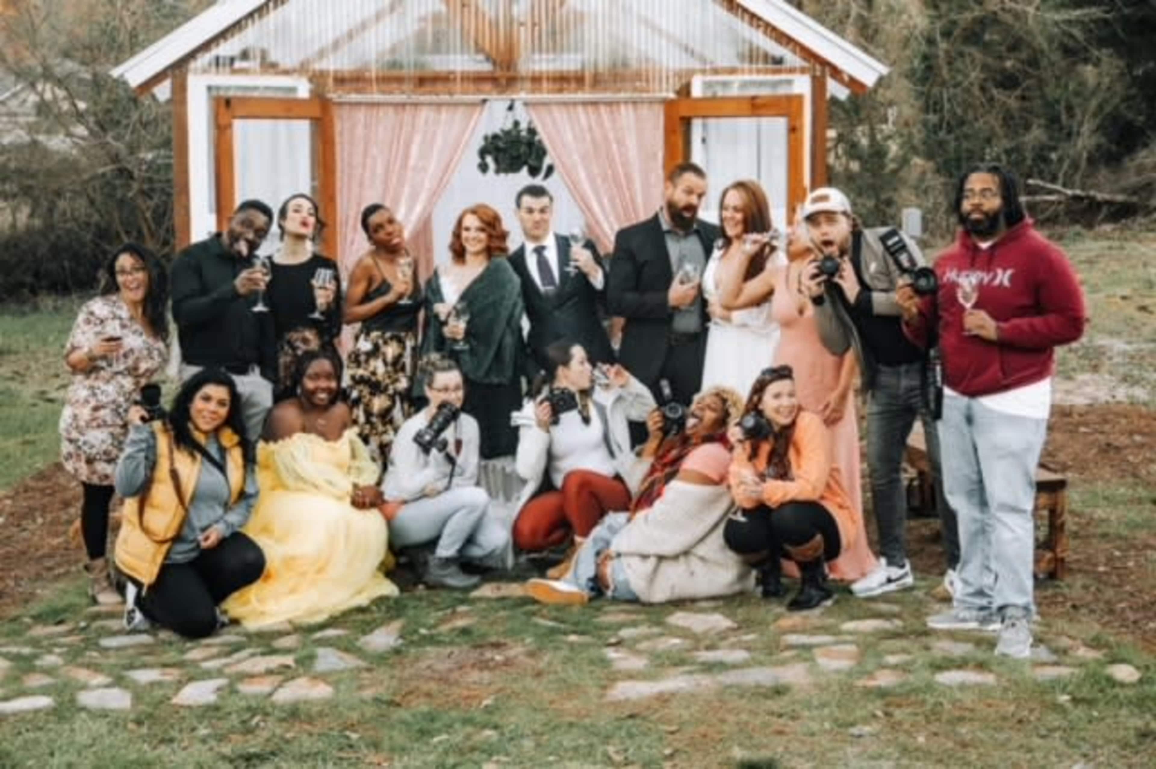 A group of people poses together in front of a decorated outdoor backdrop, with some holding drinks and cameras.