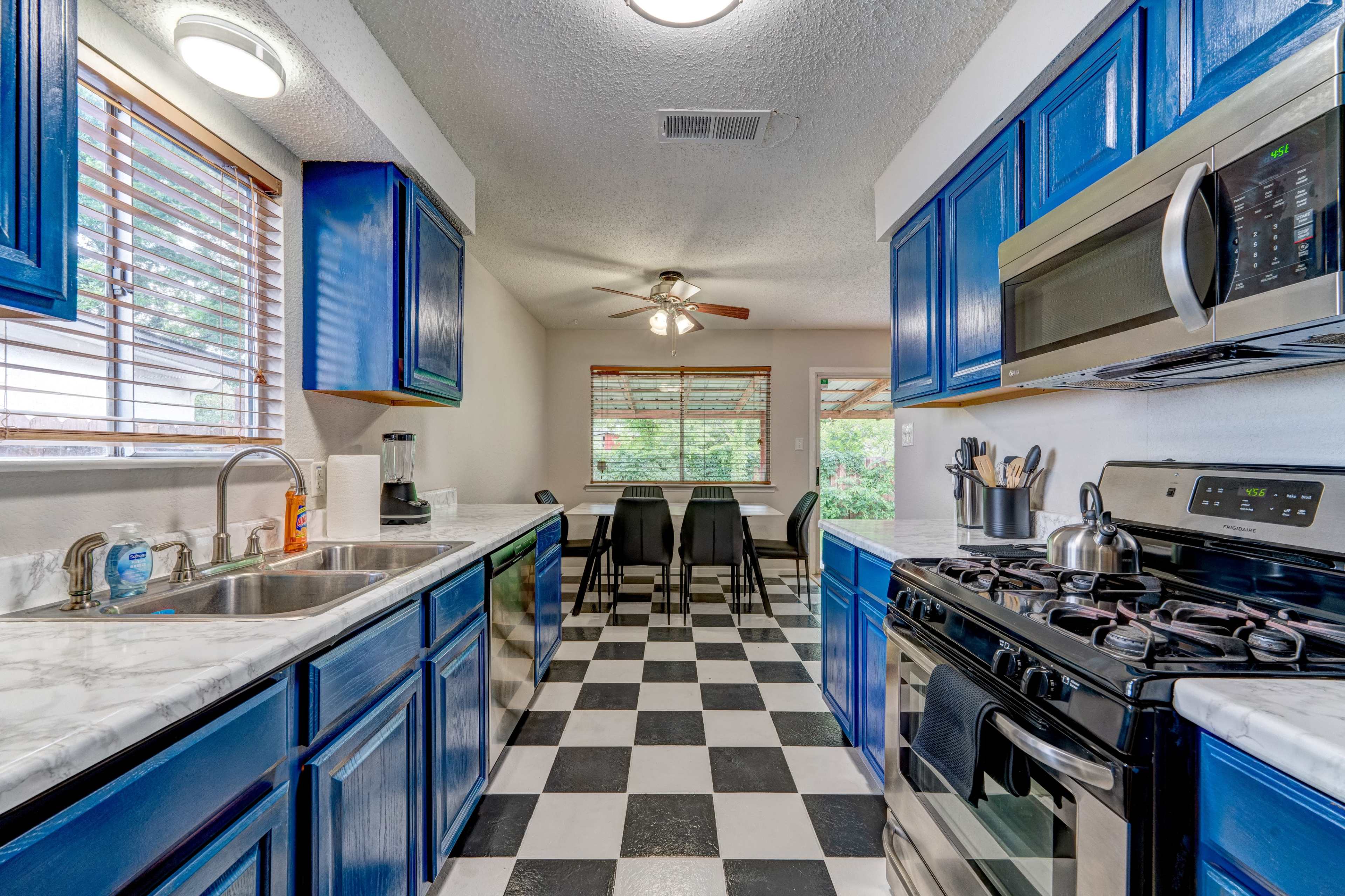 A modern kitchen features blue cabinets, a marble countertop, and a checkered black-and-white floor, with a dining area visible through the window.