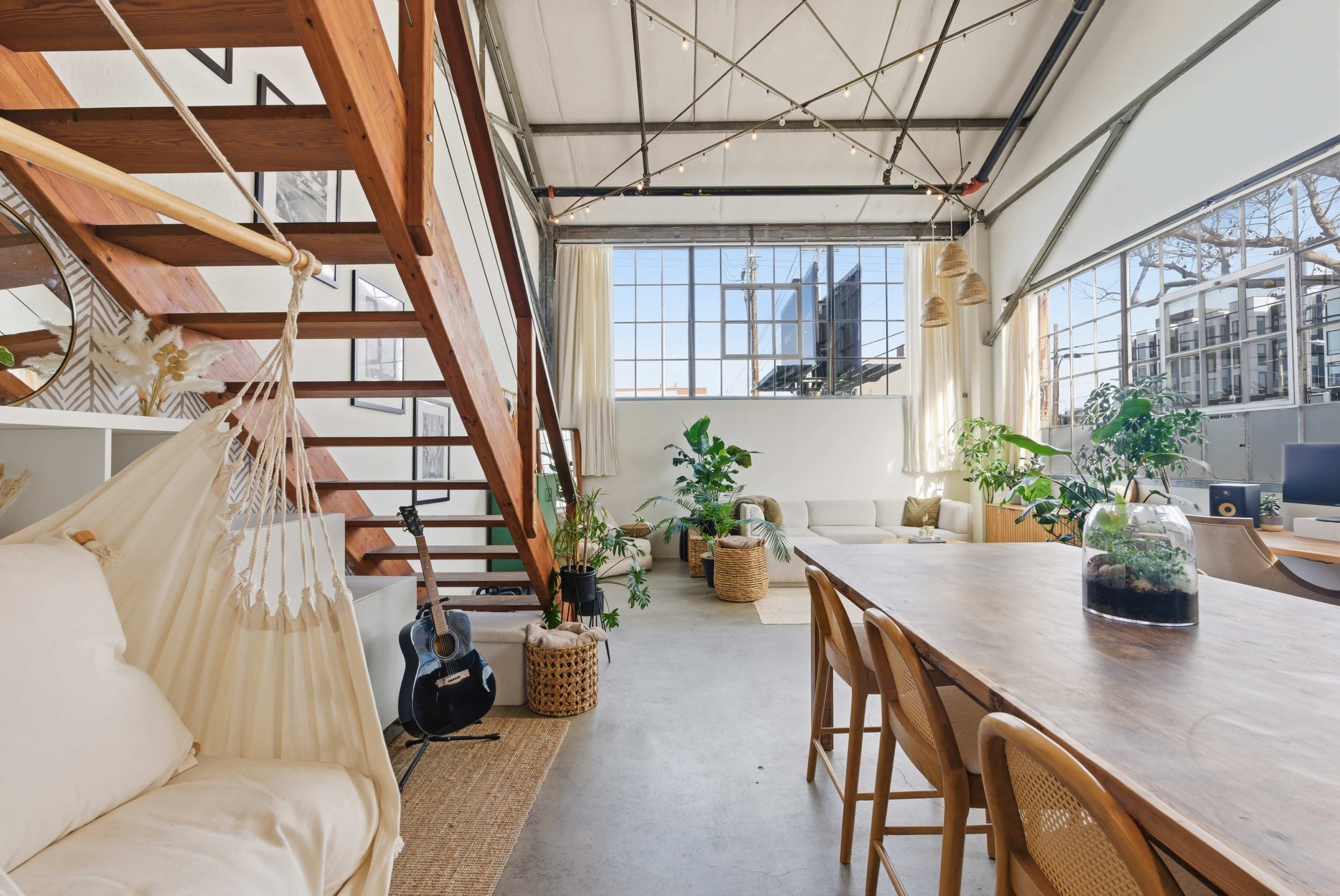 The image shows an open-plan loft with wooden stairs, a large window, plants, and a dining table framed by cozy seating areas.