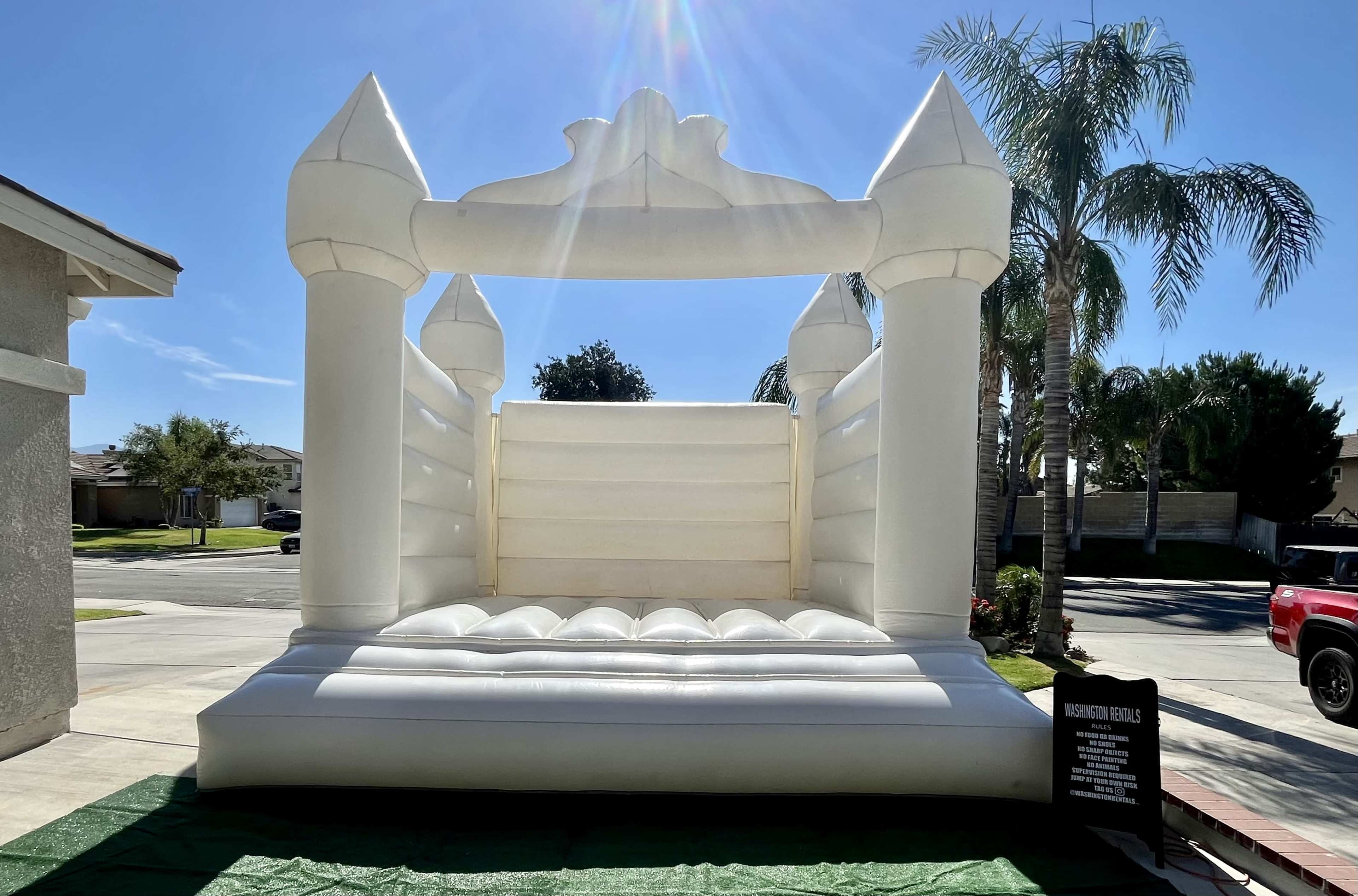 A large white inflatable bounce house stands in a front yard under a sunny sky, with palm trees visible in the background.