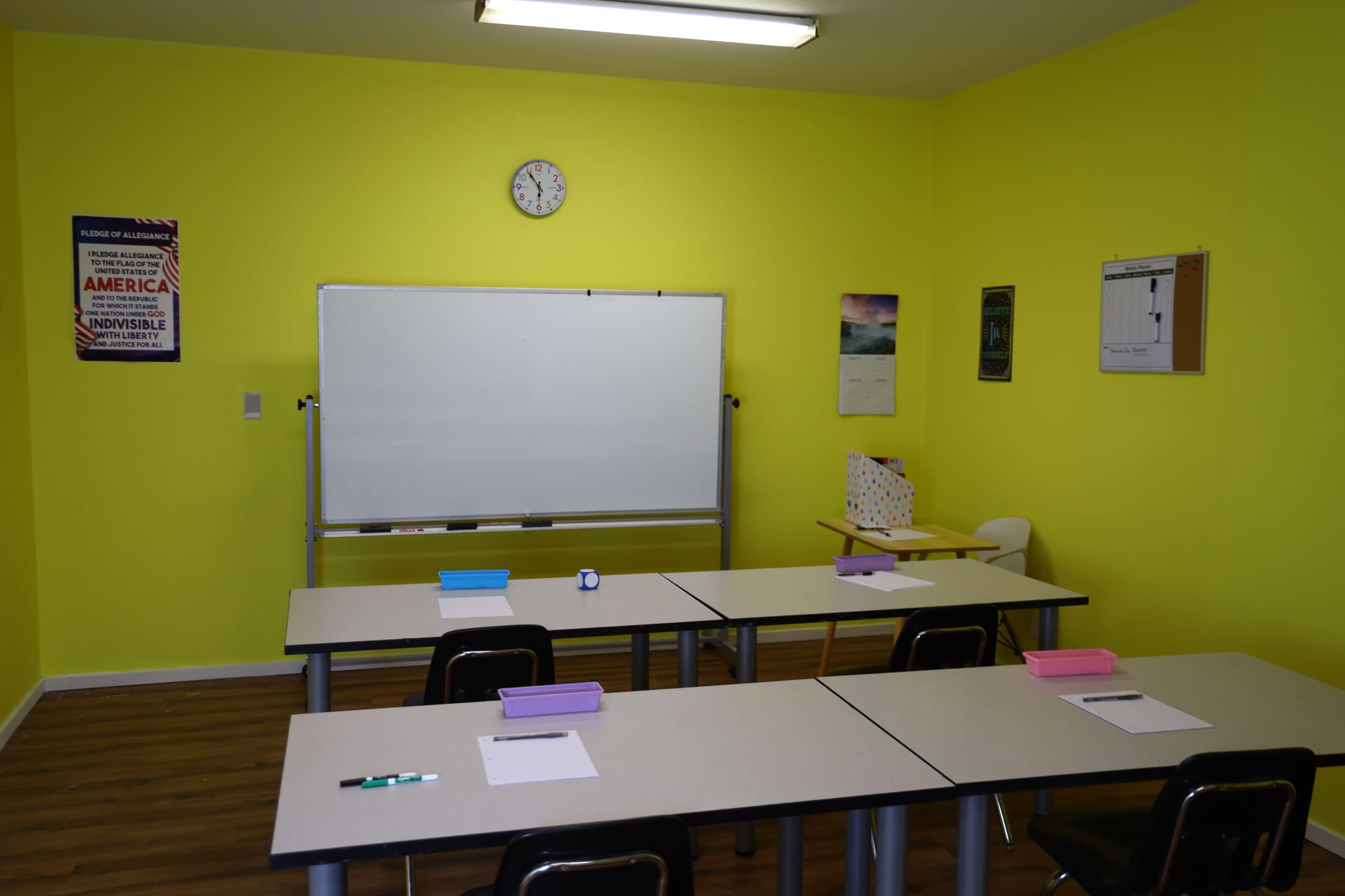 A bright yellow classroom features two tables with pink and white stationery, a whiteboard, a clock, and various posters on the walls.