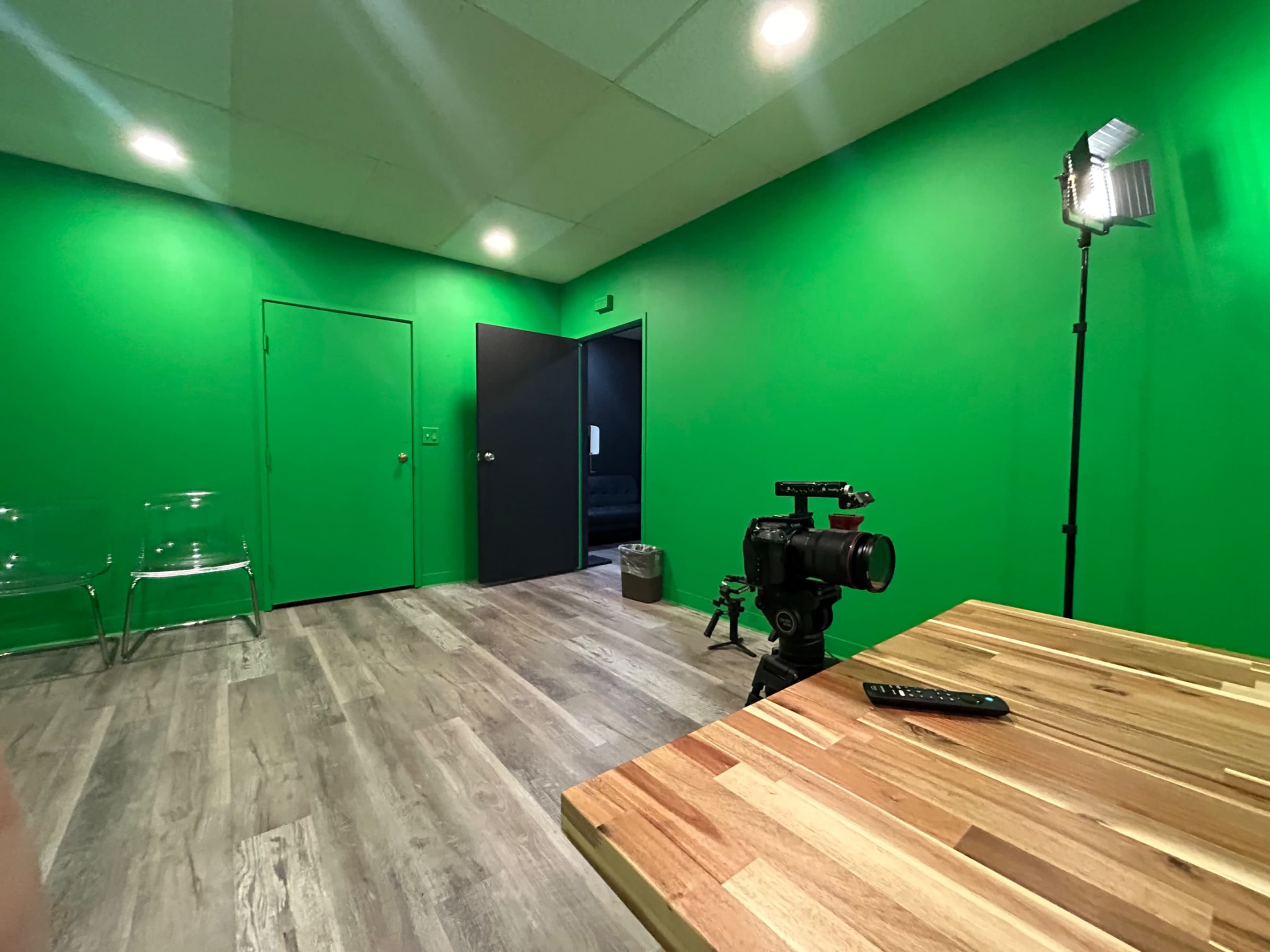 The image shows a green screen room with a camera on a tripod and a wooden table, along with clear chairs and a door leading to a dark-walled area.