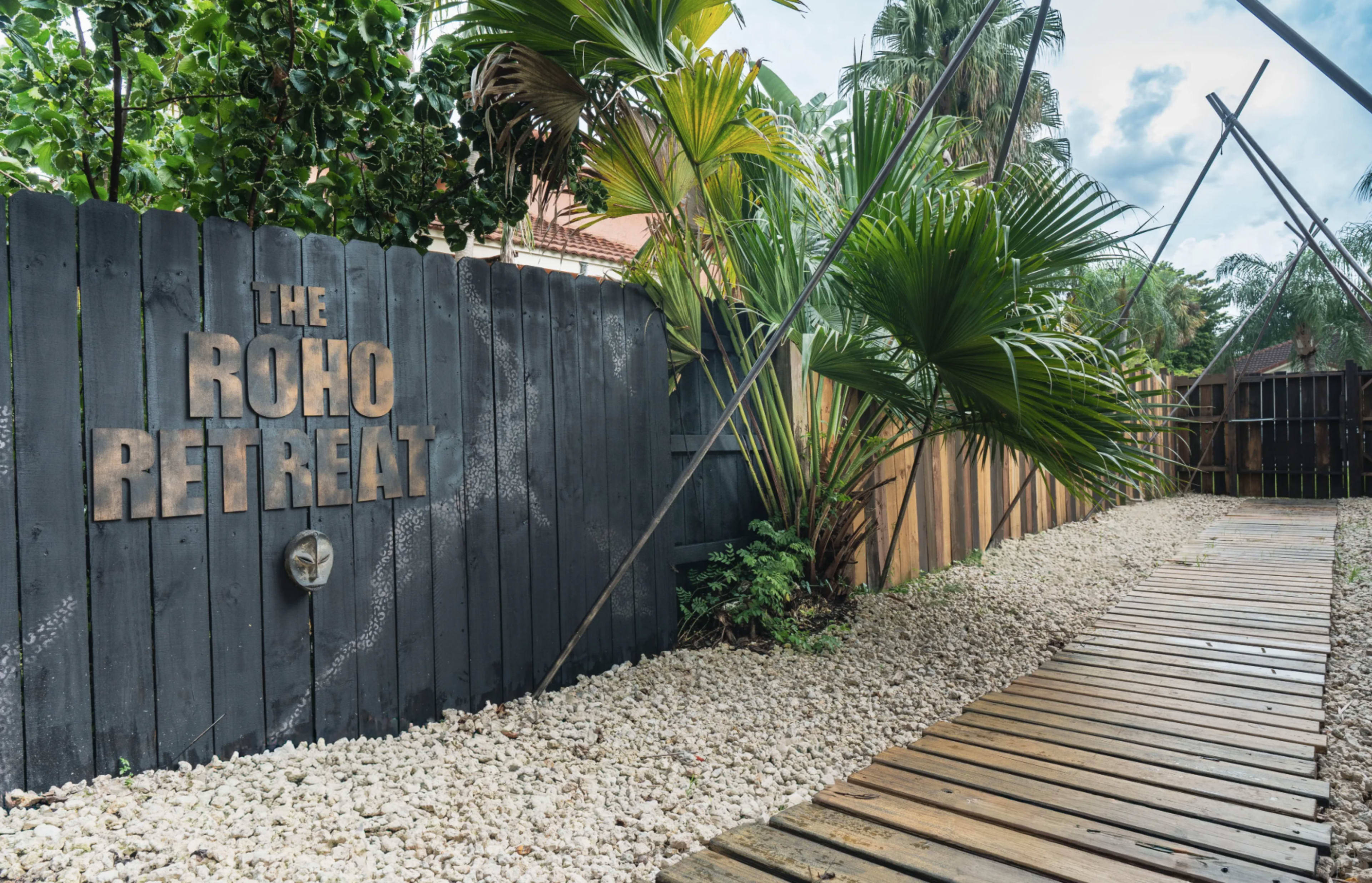 A wooden pathway leads to a fenced area marked "The Roho Retreat," surrounded by tropical plants and gravel.