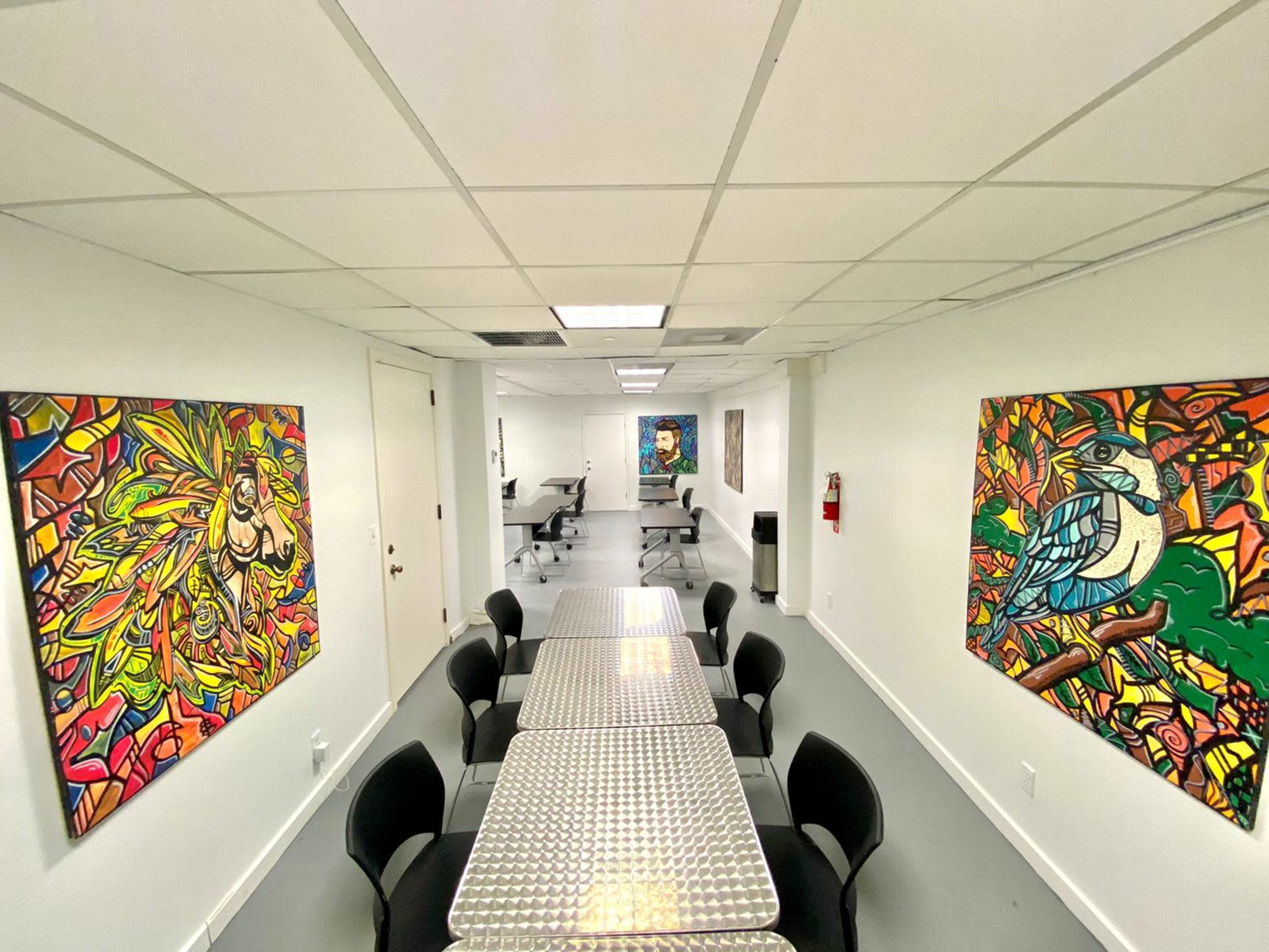 A bright, narrow room with two large, colorful paintings on the walls, several tables arranged in the center, and additional tables in the background.