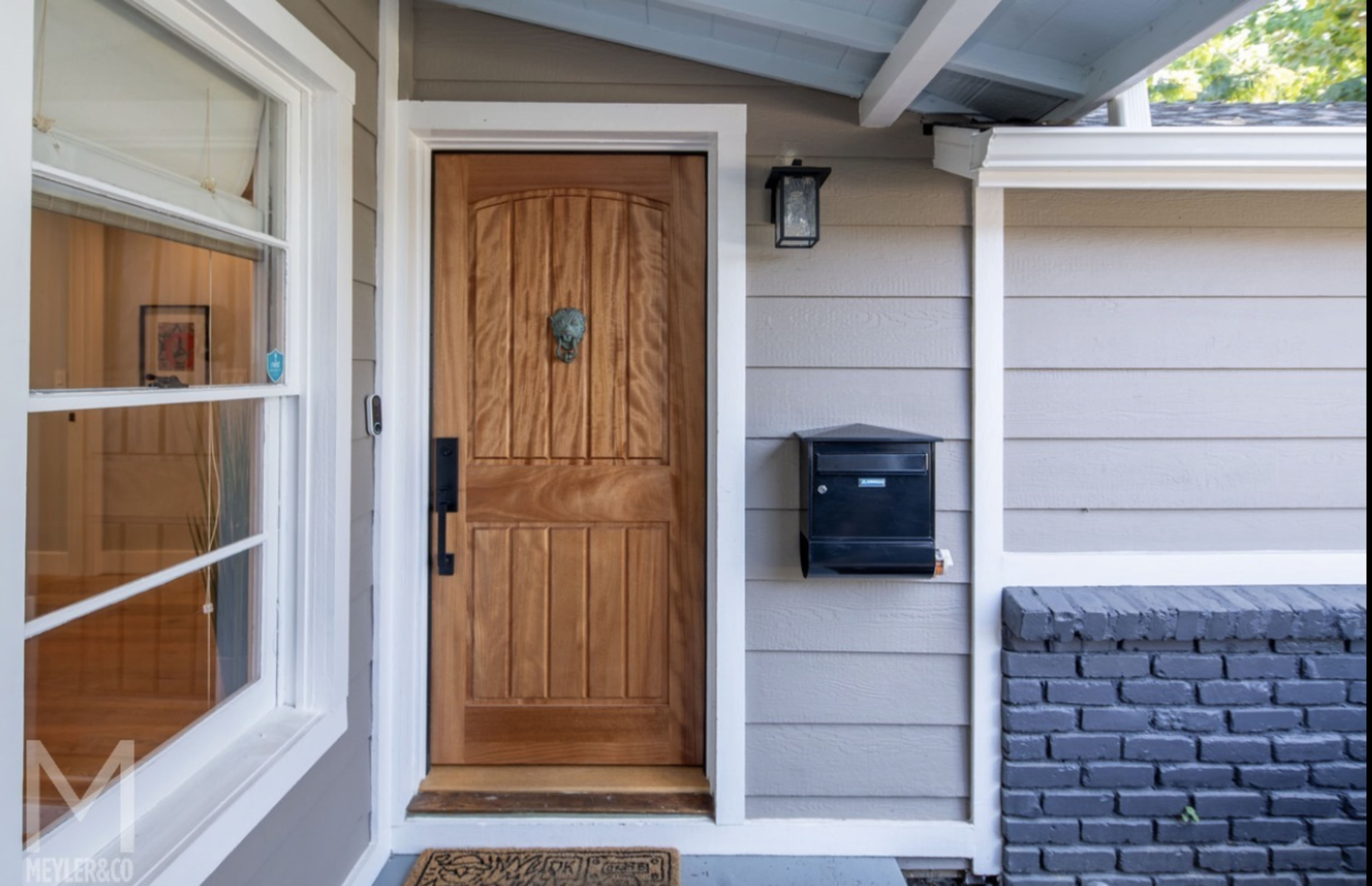 A front door with a wooden panel, a lion's head door knocker, and a black mailbox beside it on a gray house facade.