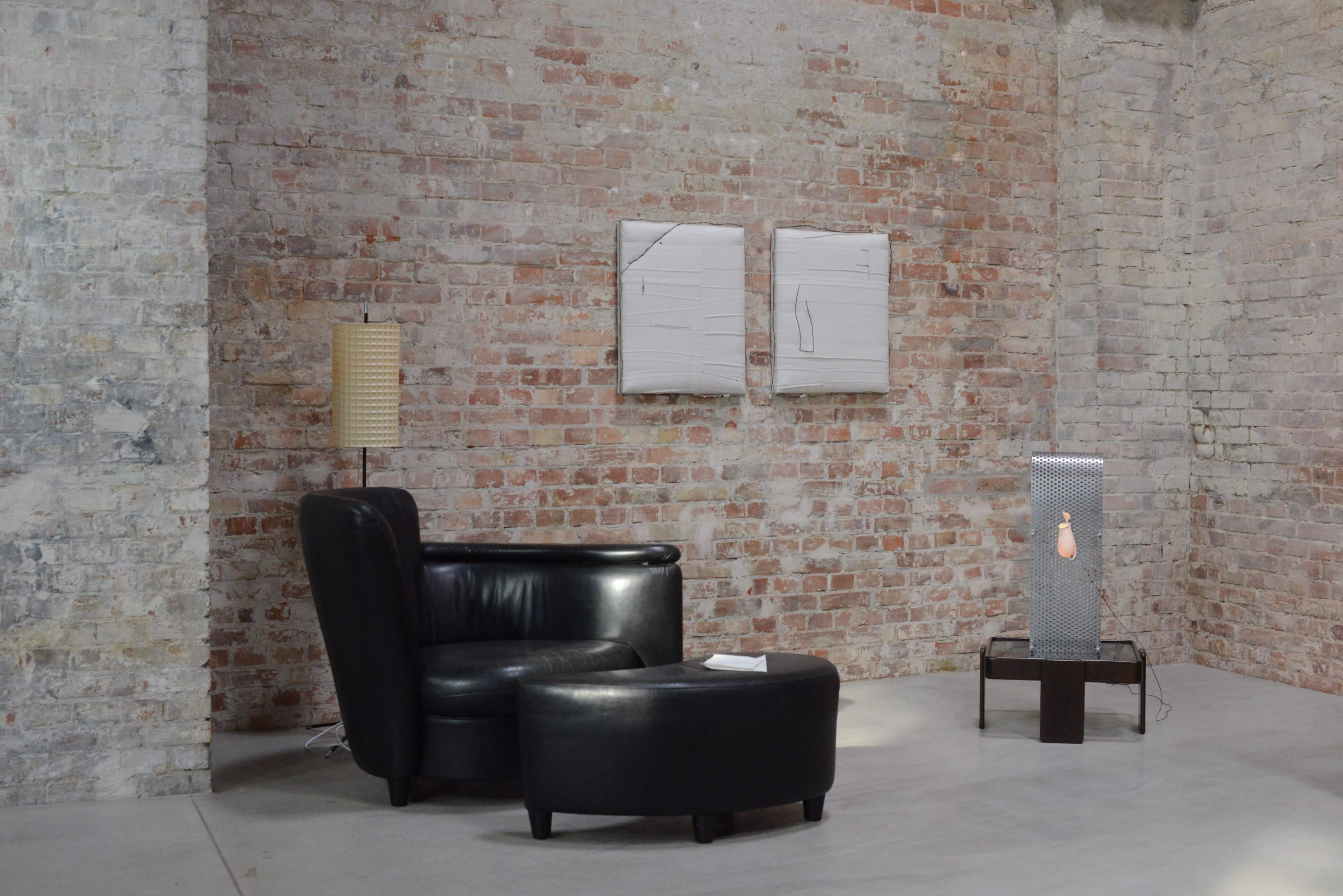 The image shows a modern room with exposed brick walls, featuring a black leather armchair and ottoman, a contemporary lamp, two abstract paintings, and a metallic sculpture on a table.